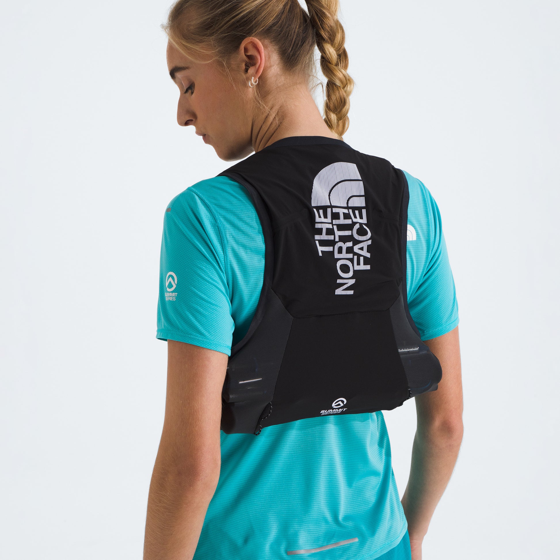 A person with a braid wears a turquoise shirt and a black "U Summit Run Vest 5" by The North Face, facing slightly away.