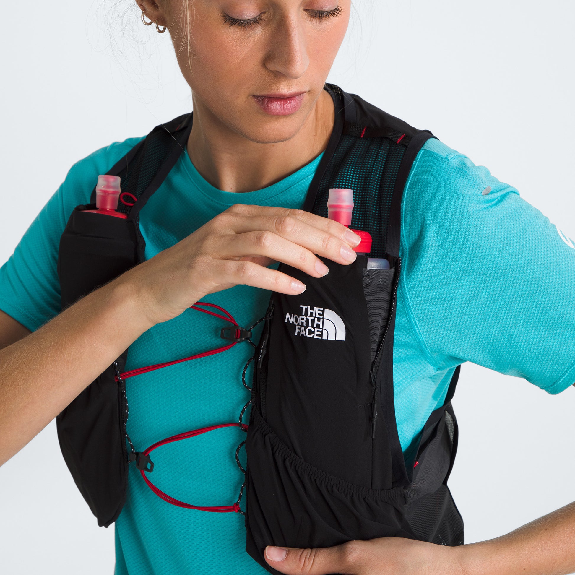 A person wearing a turquoise shirt adjusts their black The North Face U Summit Run Vest 5 with red details, featuring two small water bottles in the front pockets, preparing for race day.