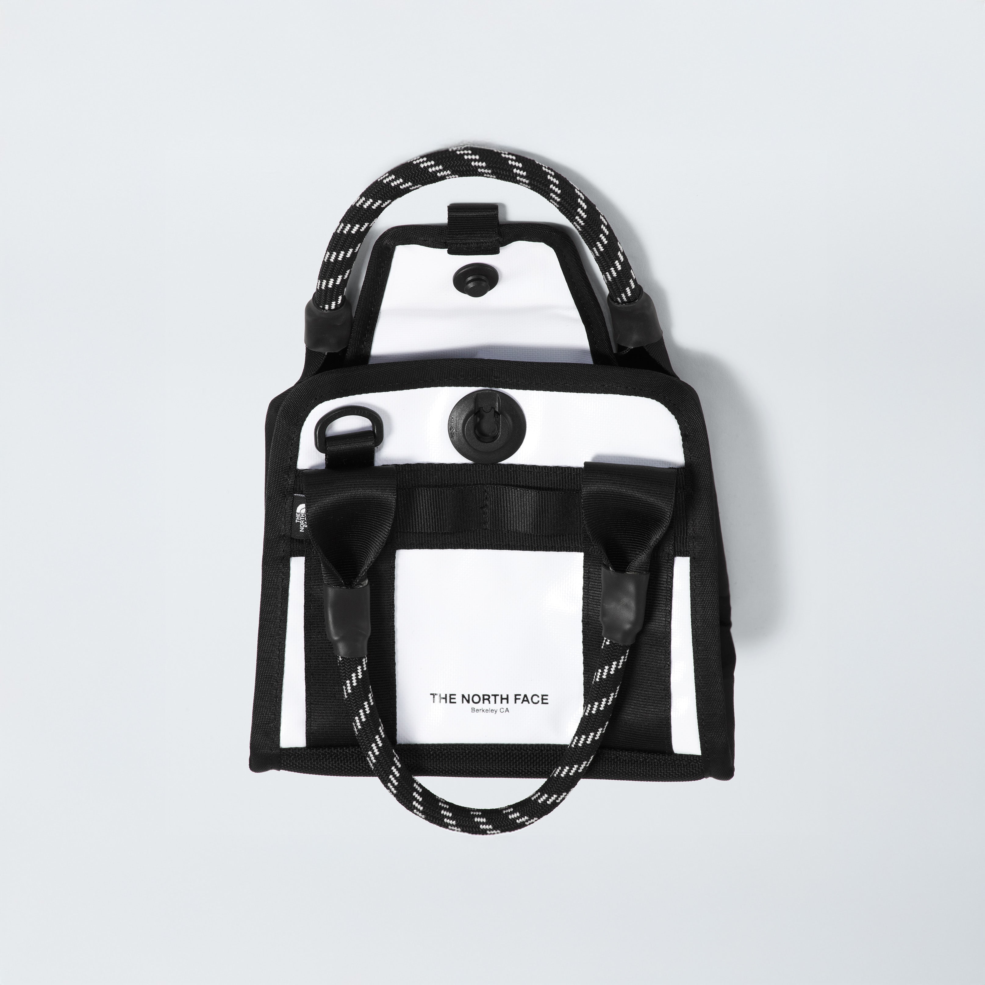 The Base Camp Mini Tote from The North Face is a sleek black and white rectangular bag made from recycled fabric, featuring rope handles, an internal organization system, a front pocket, and the iconic logo.
