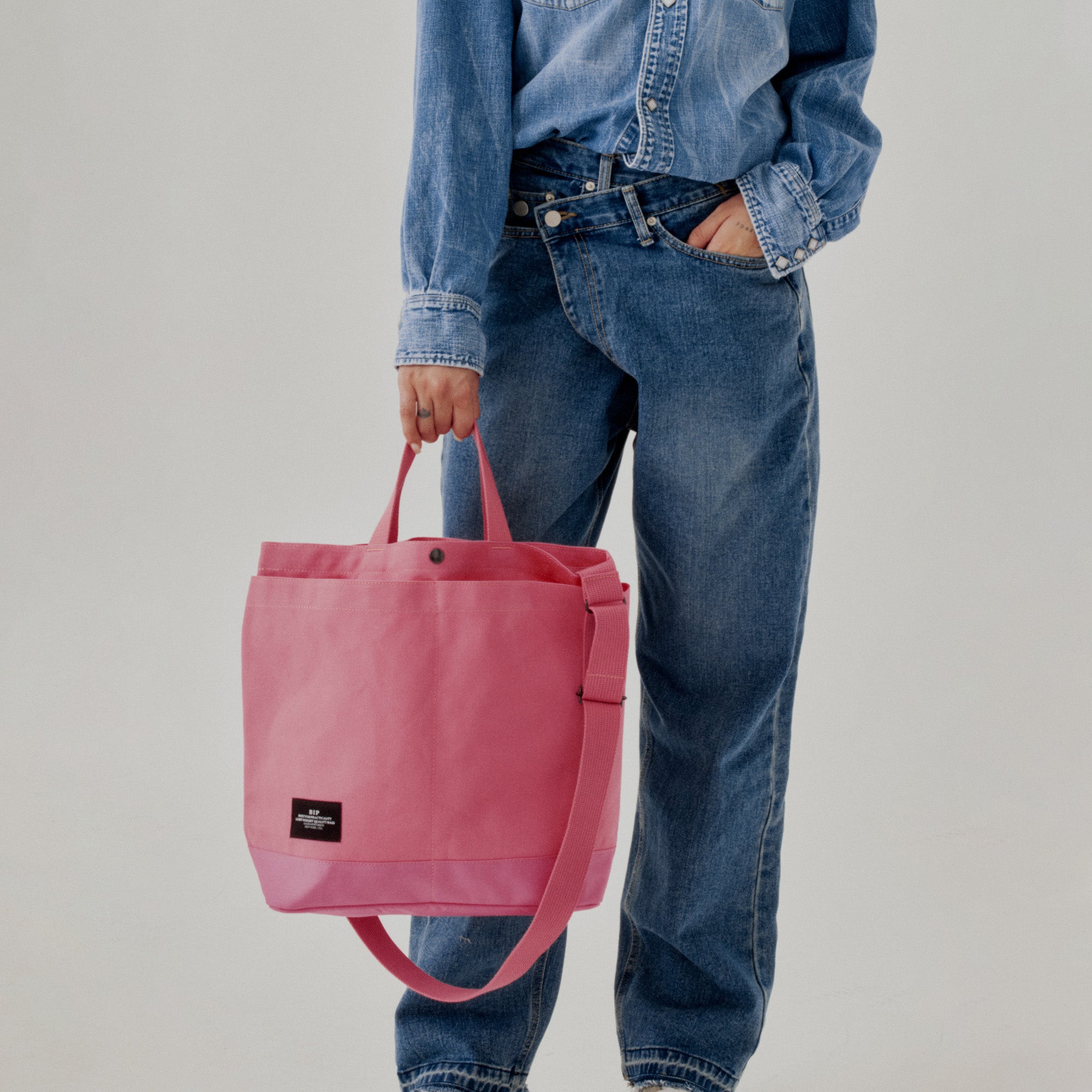 A person in a denim shirt and jeans holds the Bags in Progress New York Tote Medium, Special Edition: Candy Pink, featuring an open top and adjustable shoulder strap, against a plain background.