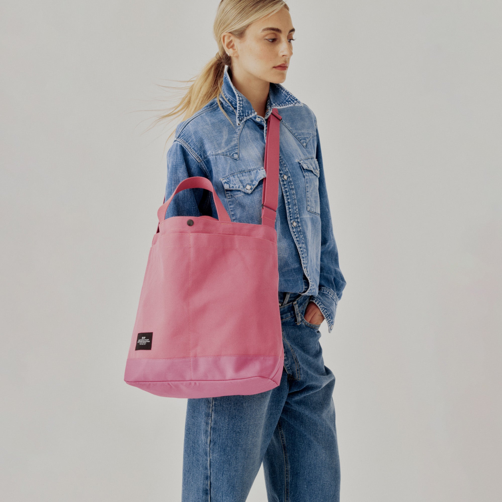 A person in a denim jacket and jeans holds a Bags in Progress New York Tote Medium, Special Edition: Candy Pink, featuring an adjustable shoulder strap, open top, and black label.