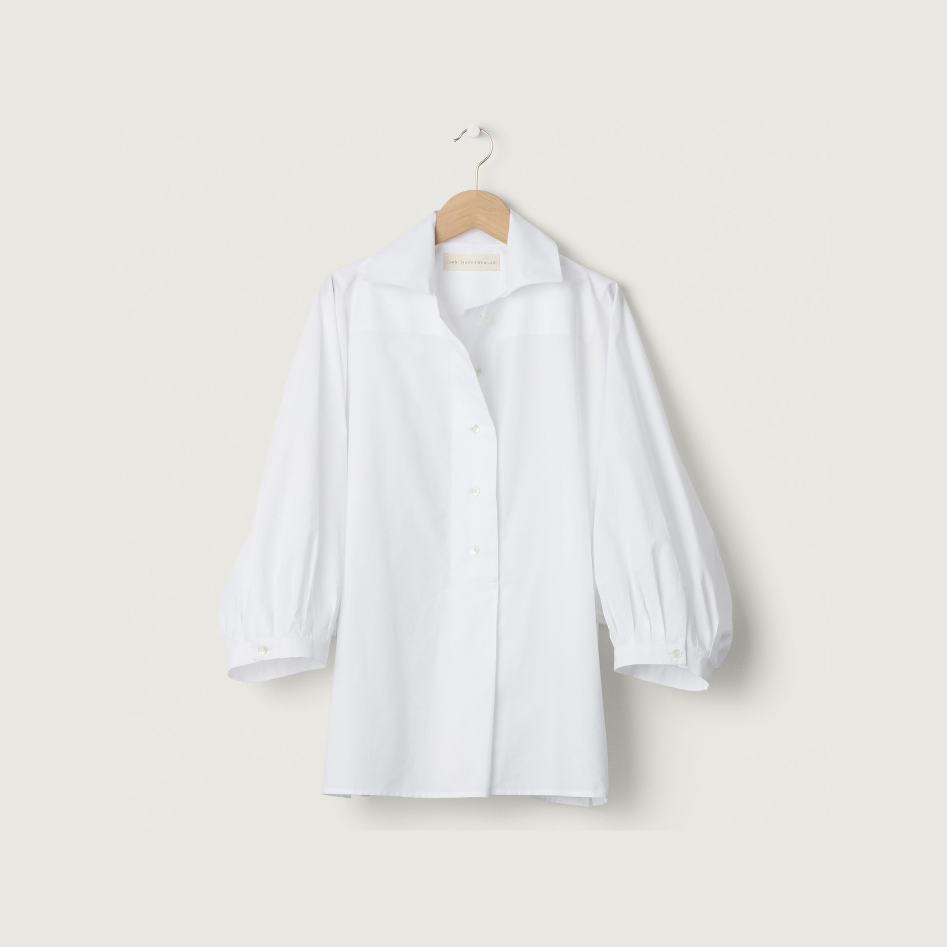 The Nina Shirt in Poplin White by Jan Machenhauer, featuring long slightly puffed sleeves, hangs on a wooden hanger against a plain light background.