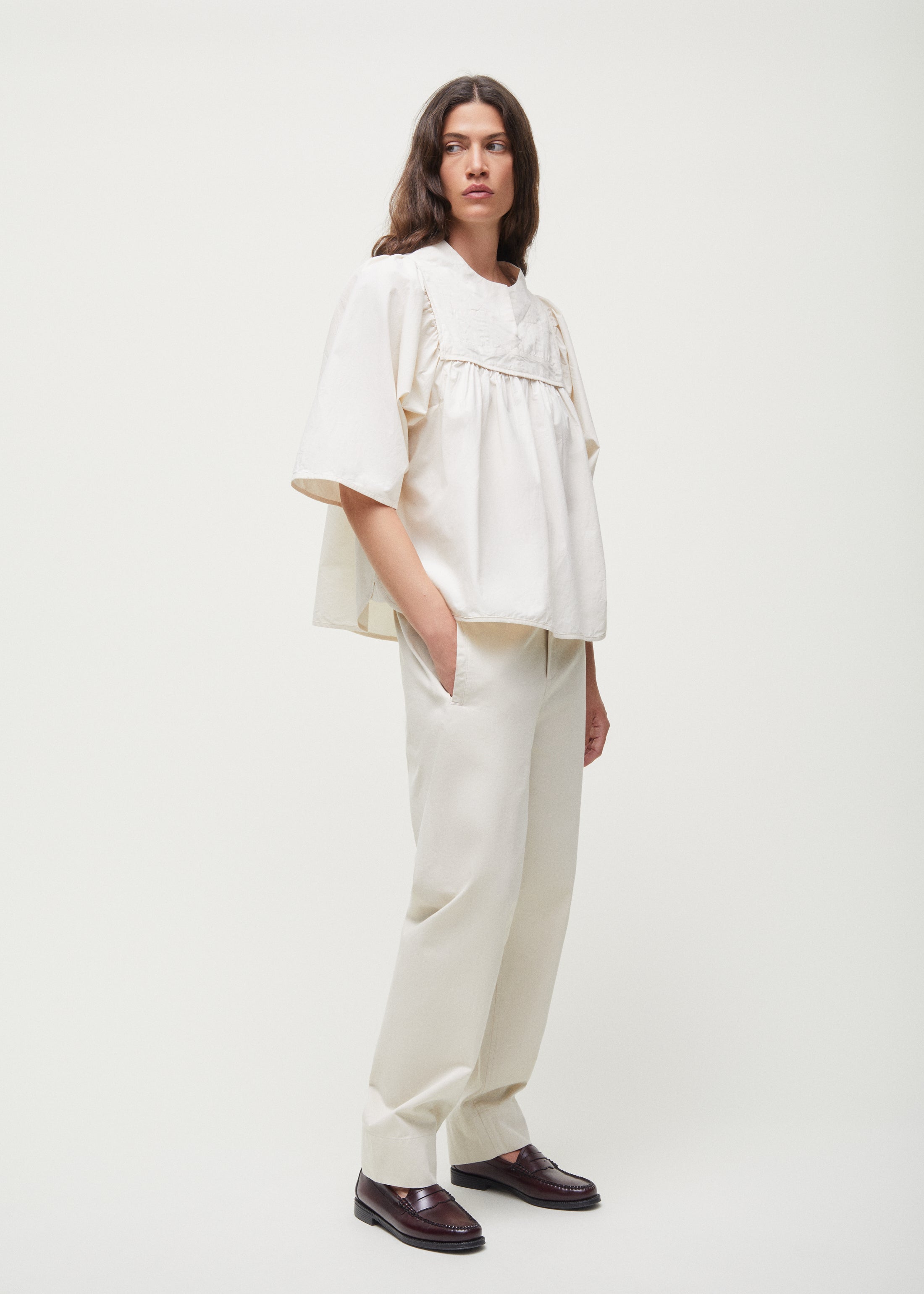 A person with long hair, wearing the loose-fitting Nomi Blouse in beige, crafted by Aiayu from regenerative cotton, paired with matching trousers, stands against a plain background with hands in pockets.