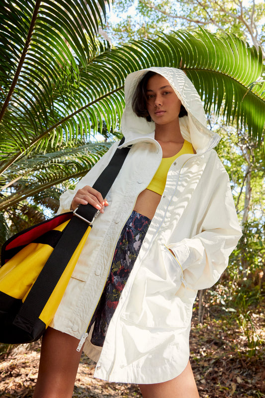 A young woman in a stylish Singin in the Rain Jacket by Free People Movement and colorful outfit stands confidently under palm leaves in a sunlit forest.