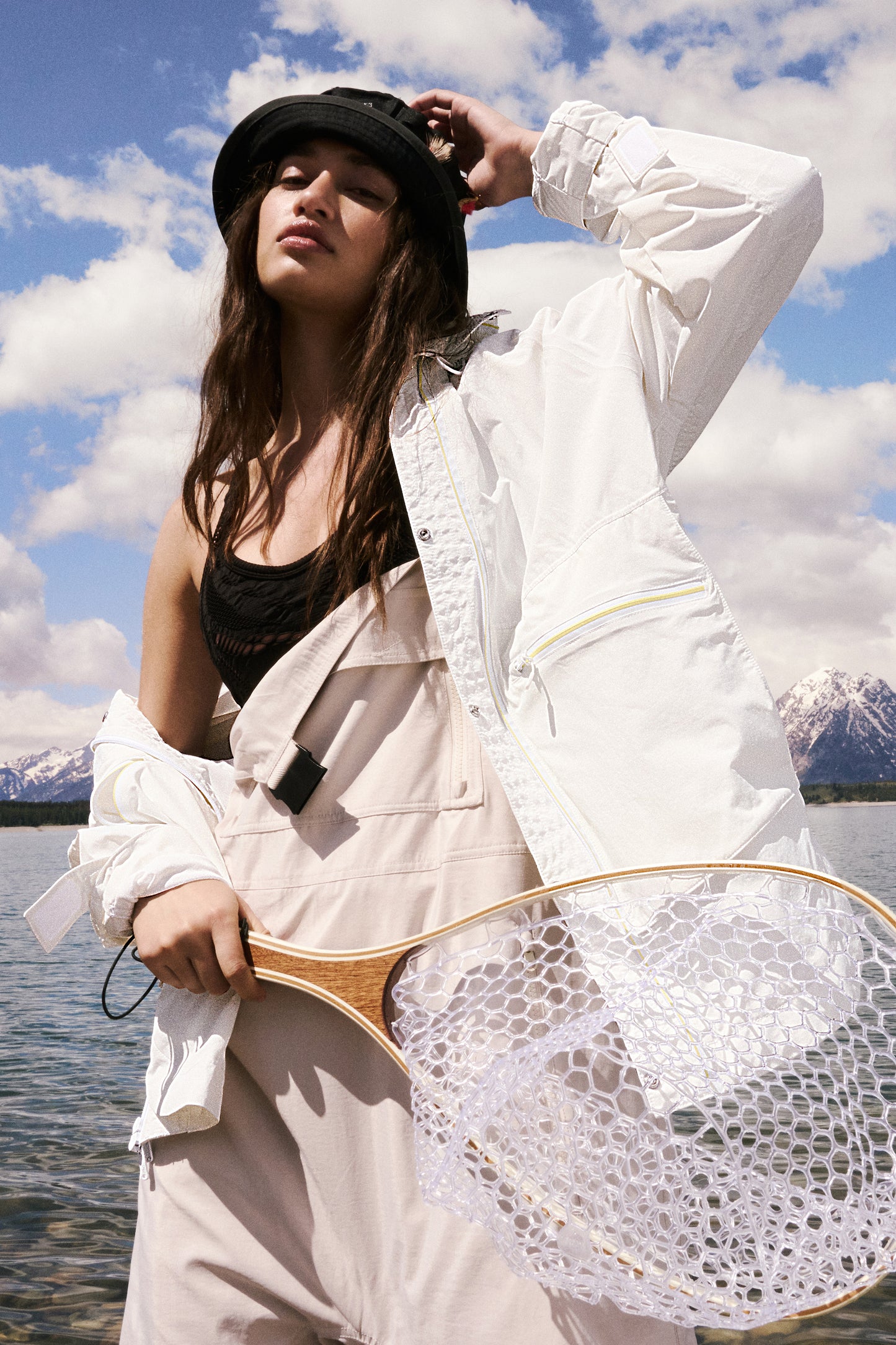 A woman wearing a Singin in the Rain Jacket by Free People Movement and holding a fishing net stands by a lake, adjusting her black hat, with snowy mountains in the background.