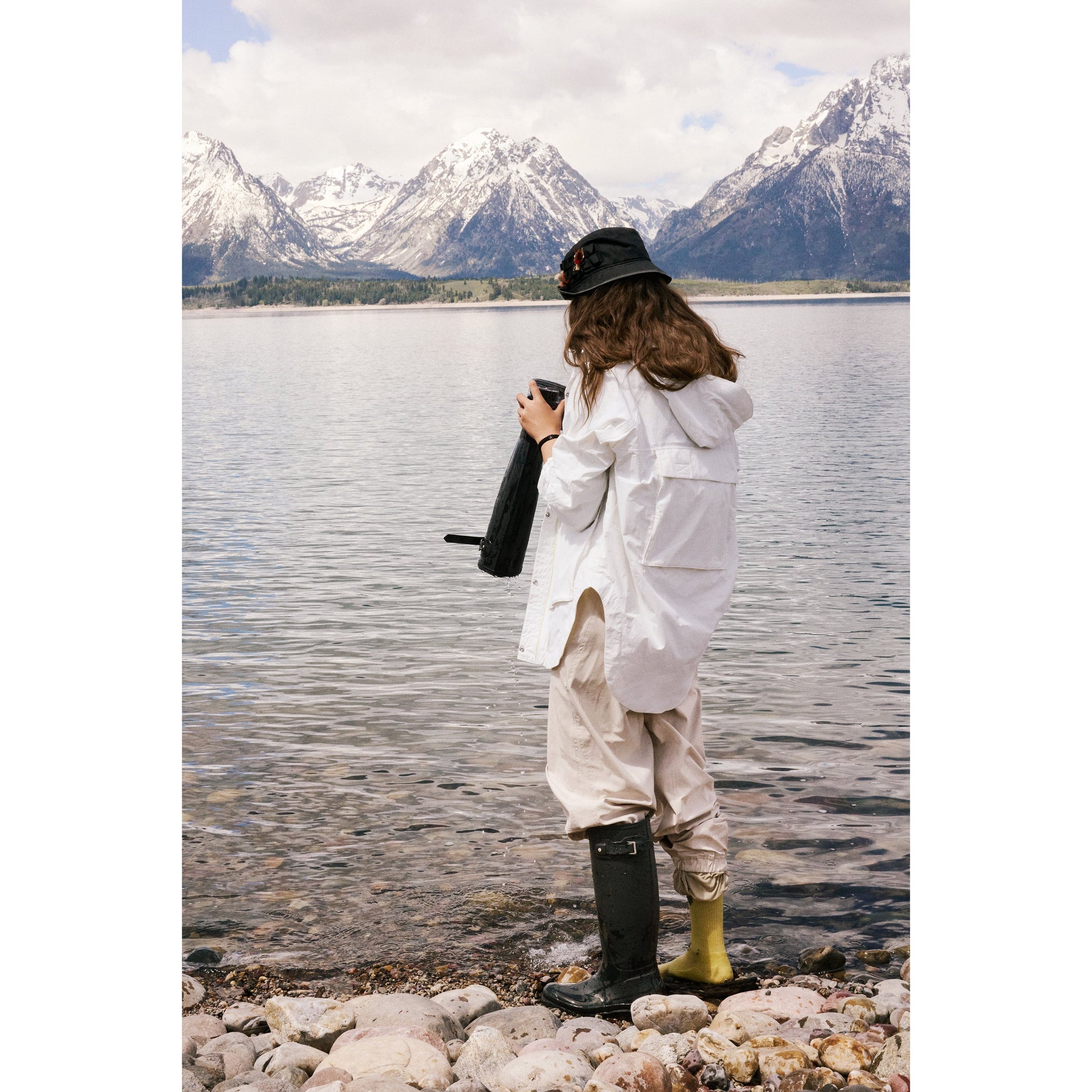Woman standing by a lake, looking at her phone, wearing a Free People Movement Singin in the Rain Jacket in Painted White and black hat, with snowy mountains in the background.