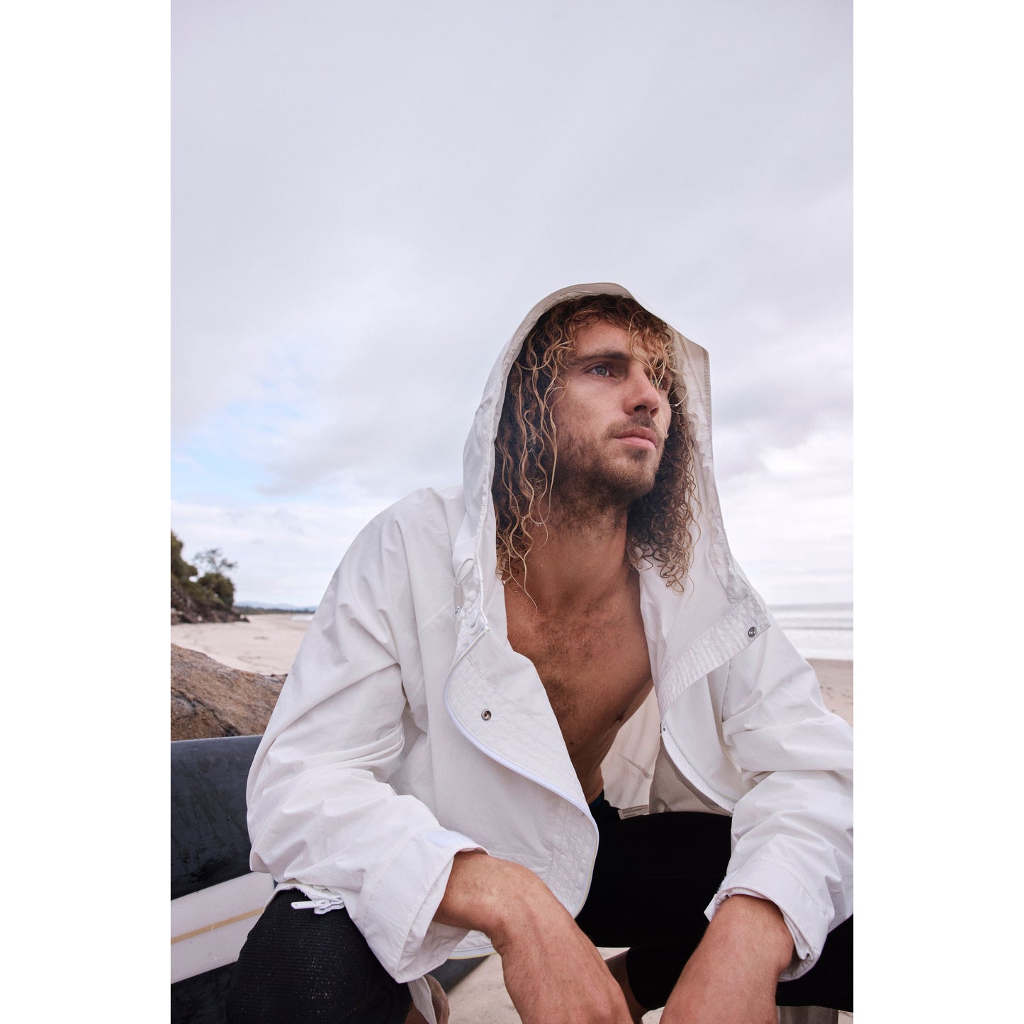 Man with curly hair wearing a Free People Movement Singin in the Rain Jacket in Painted White with hood, sitting on a beach on an overcast day, looking thoughtfully into the distance.