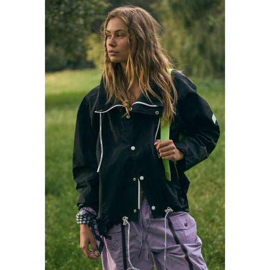 A young woman in a casual Rain & Shine Jacket in black by Free People Movement and lilac pants walks in a green park, looking thoughtfully to the side.