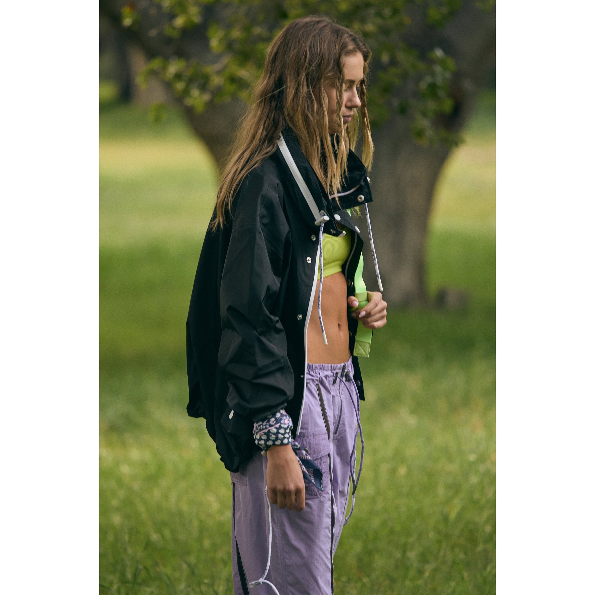 A young woman standing in a grassy field, wearing a Free People Movement Rain & Shine Jacket in black, lime green crop top, and purple track pants.