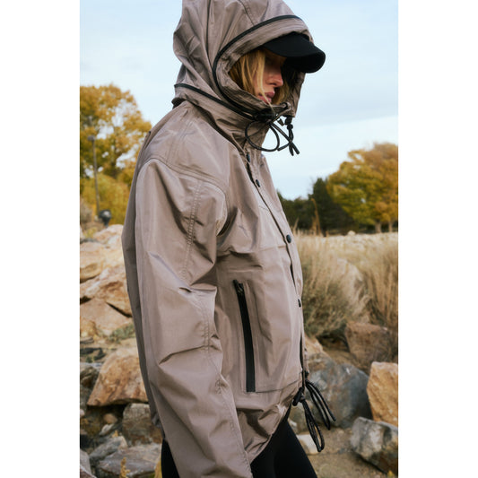 Woman in a Rain & Shine Jacket by Nordic Trail standing outdoors with a serene autumnal background, featuring rocky terrain and dried grass.