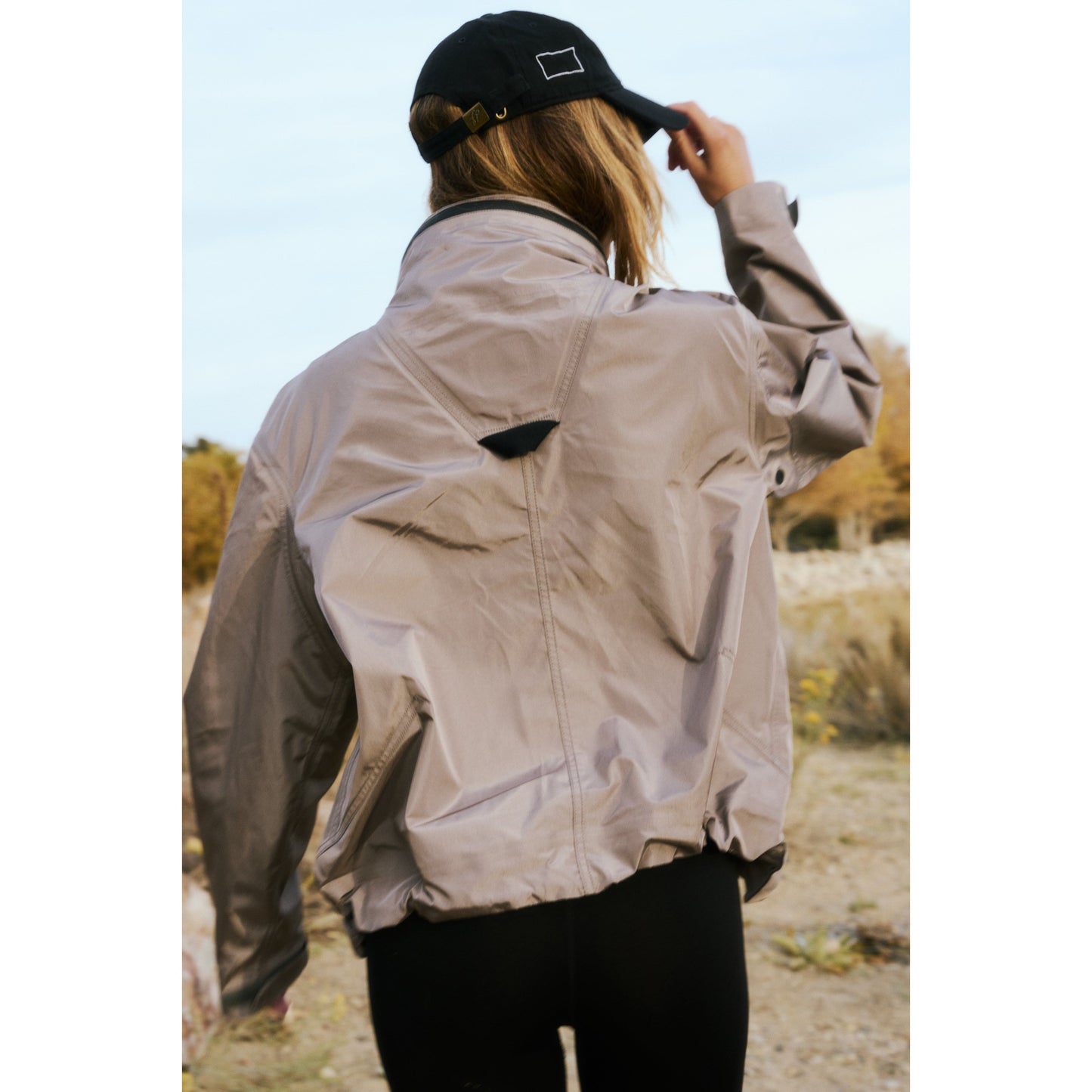 Woman from behind holding a black cap, wearing a Rain & Shine Jacket from Free People Movement, in a natural outdoor setting.