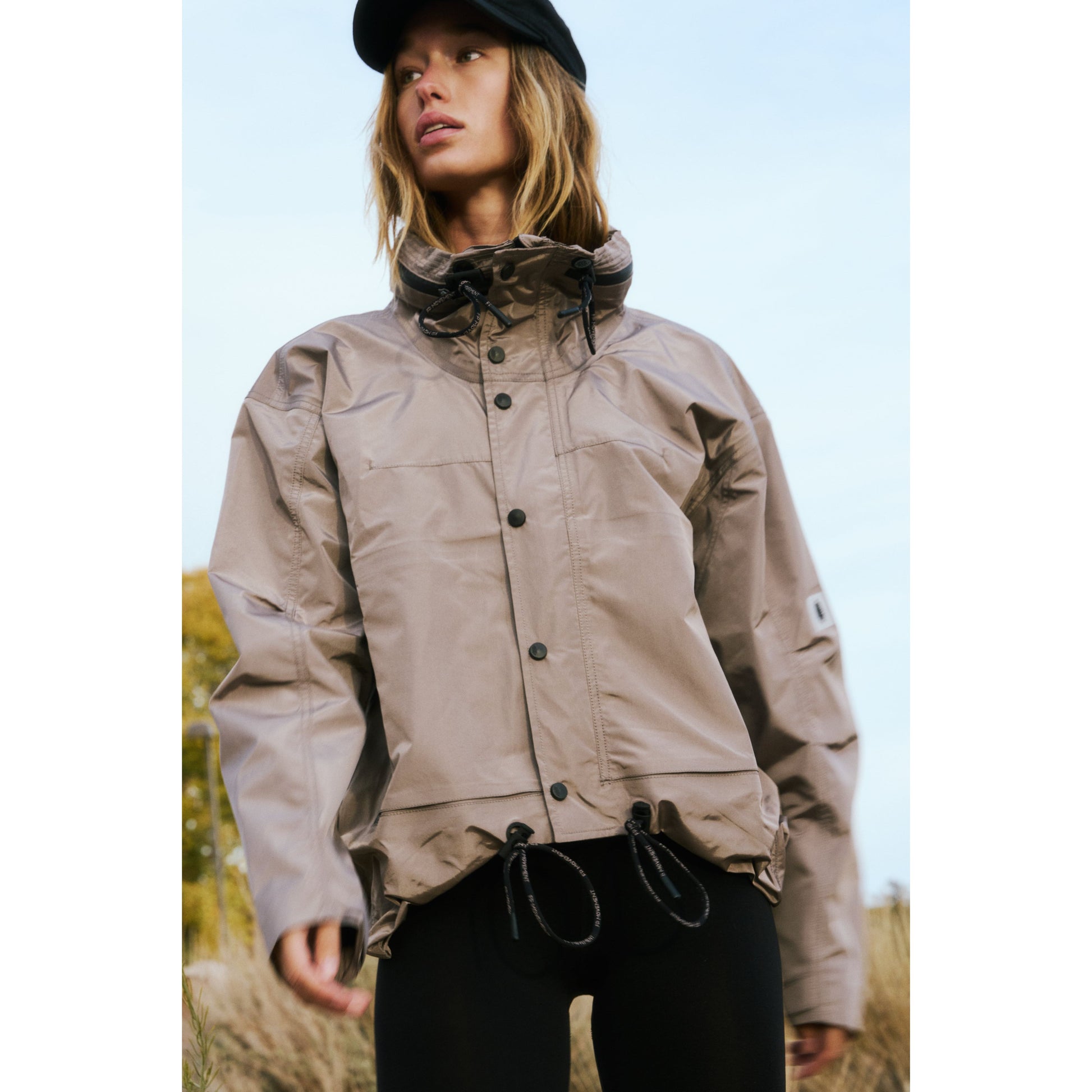 A young person wears a Rain & Shine Jacket and black pants, standing outdoors with a focused look, against a blurred natural background. Brand Name: Free People Movement