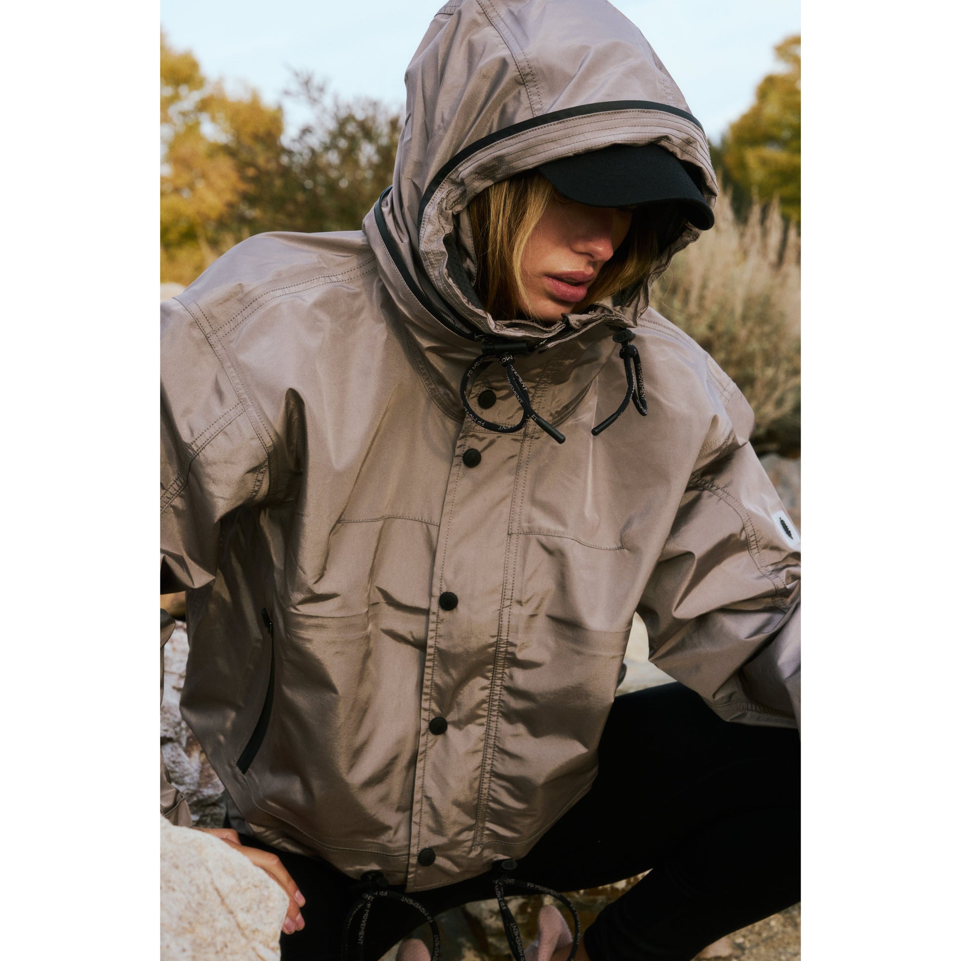 A person in a hooded beige Rain & Shine Jacket by Nordic Trail crouching among rocks, partially covering their face with the hood.