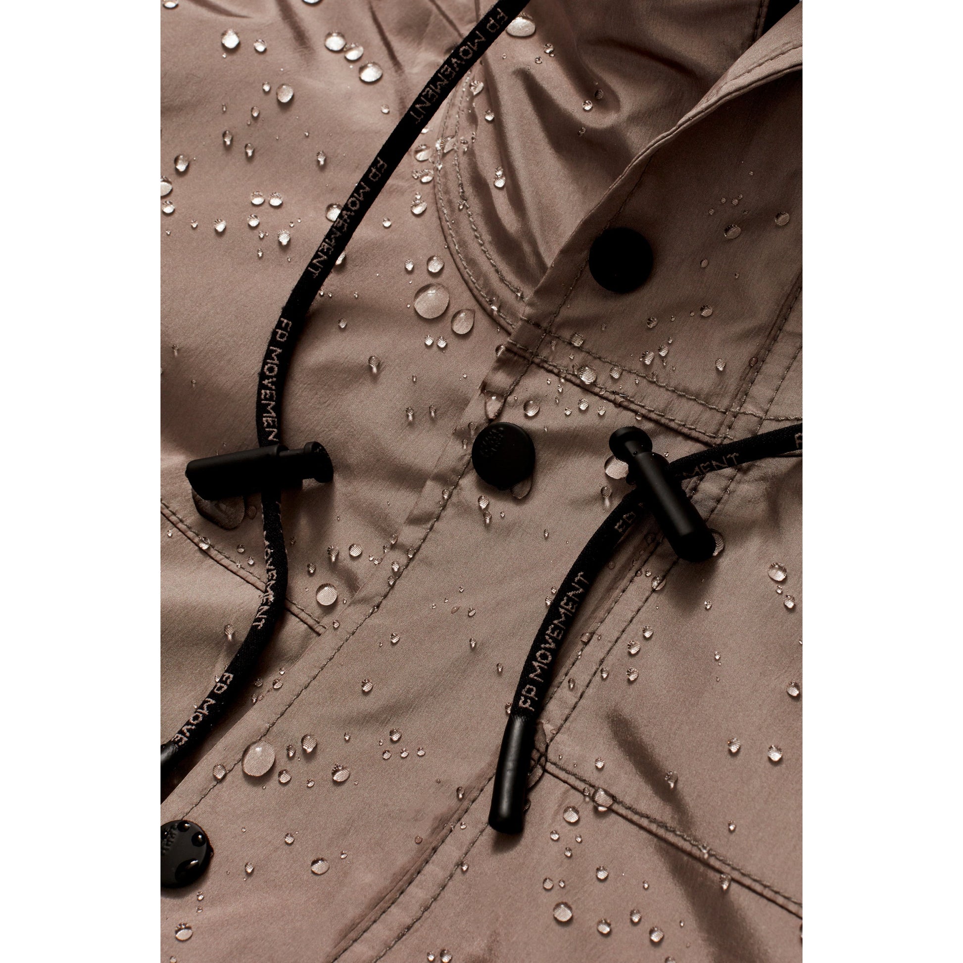Close-up of a beige Rain & Shine Jacket by Nordic Trail with water droplets on the surface, featuring visible Free People Movement brand labels and black drawstrings.