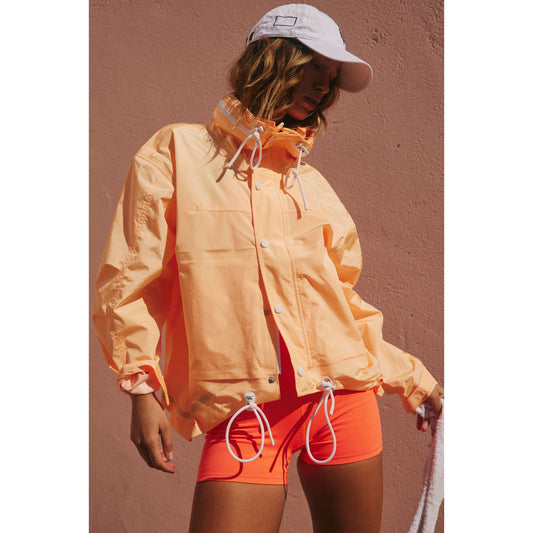 Woman in a Morning Sun Rain & Shine Jacket and orange shorts standing against a pink wall, wearing a white cap, partially in sunlight.