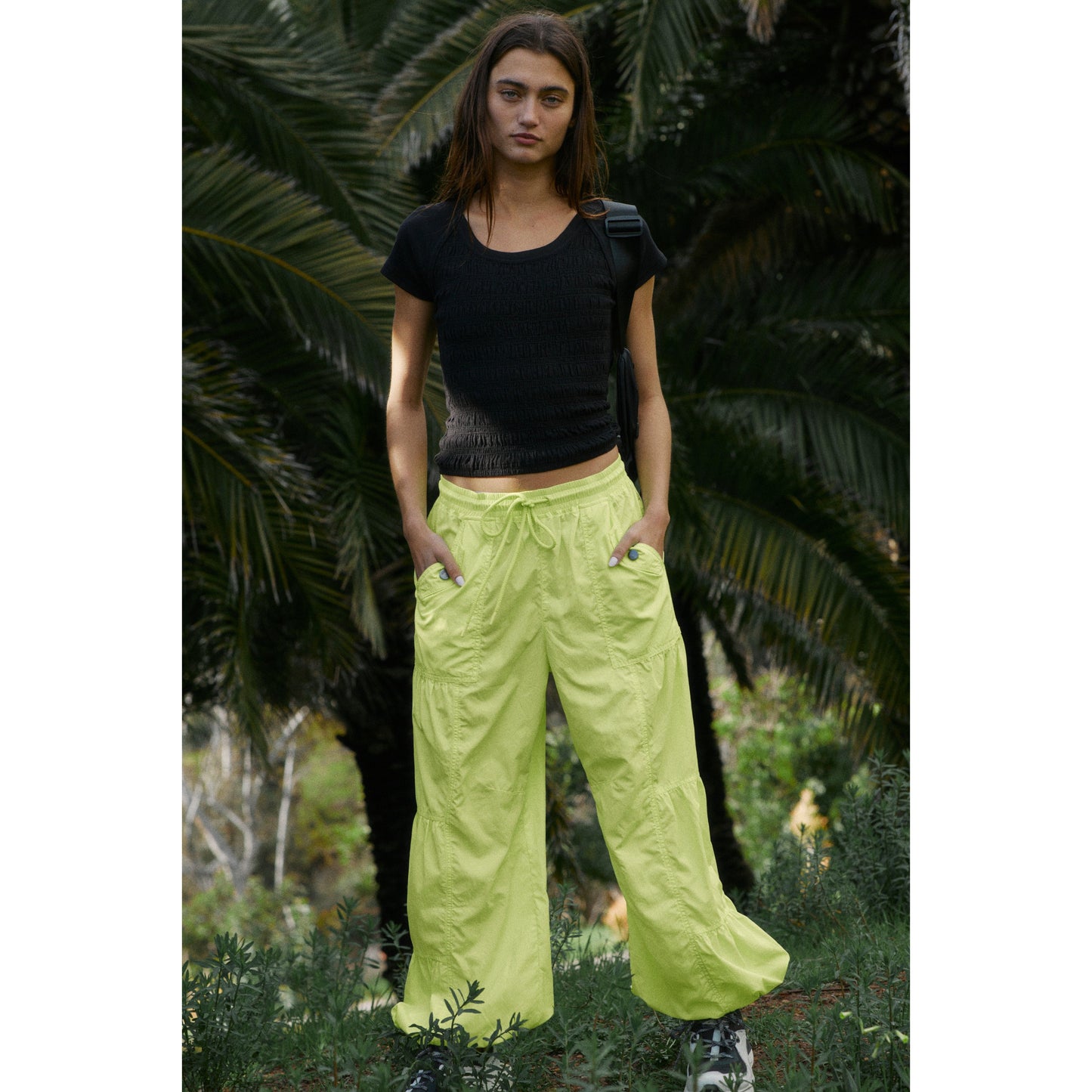 A woman with shoulder-length brown hair wearing a black top and Free People Movement's Set Me Free Pant in Lime stands in a garden, gazing directly at the camera.