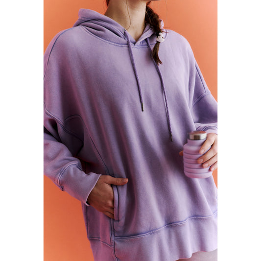 A person in a cozy Sprint To the Finish Hoodie in Vivid Lilac by Free People Movement standing against an orange background, holding a reusable drink container.
