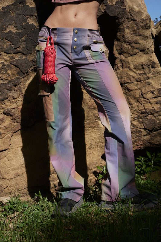 A person wearing Free People Movement's Printed Cascade Flare trousers in Galaxy Gradient and holding a coiled red rope stands against a rock background, focusing on the midsection.