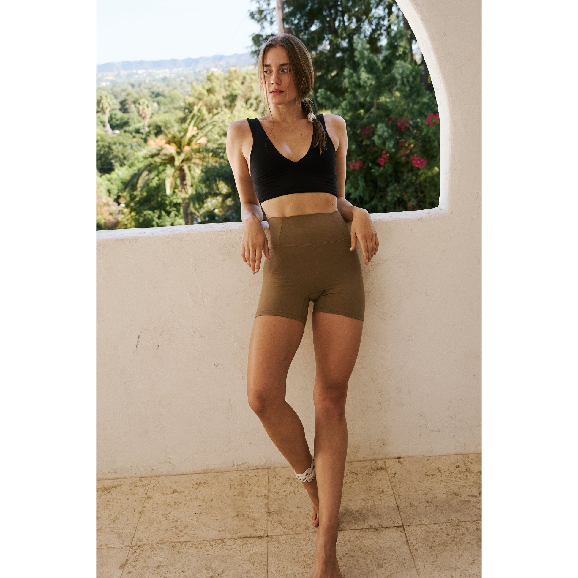 A woman in a black sports bra and Free People Movement's Moonrock Never Better Bike Shorts stands confidently on a balcony with lush greenery in the background.