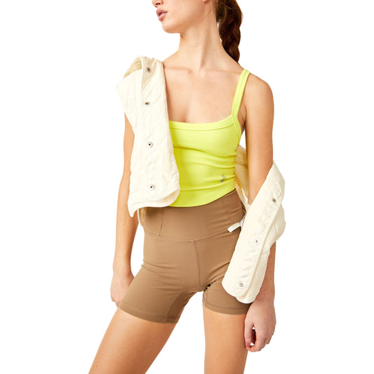 A woman wearing a Free People Movement All Clear Cami Solid in Highlighter Yellow, beige shorts, and holding a white jacket draped over her shoulder.