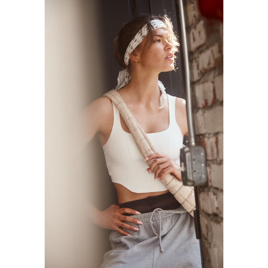 A woman in a Free People Movement white Strong Core Corset Cami and gray sweatpants leaning against a brick wall, looking out of a sunlit window.