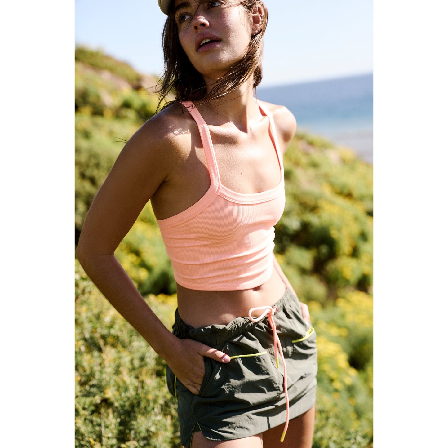 Young woman in a pink tank top and green shorts standing on a sunny hillside with the ocean in the background wearing a Free People Movement Outskirts Skort in Olive Combo.