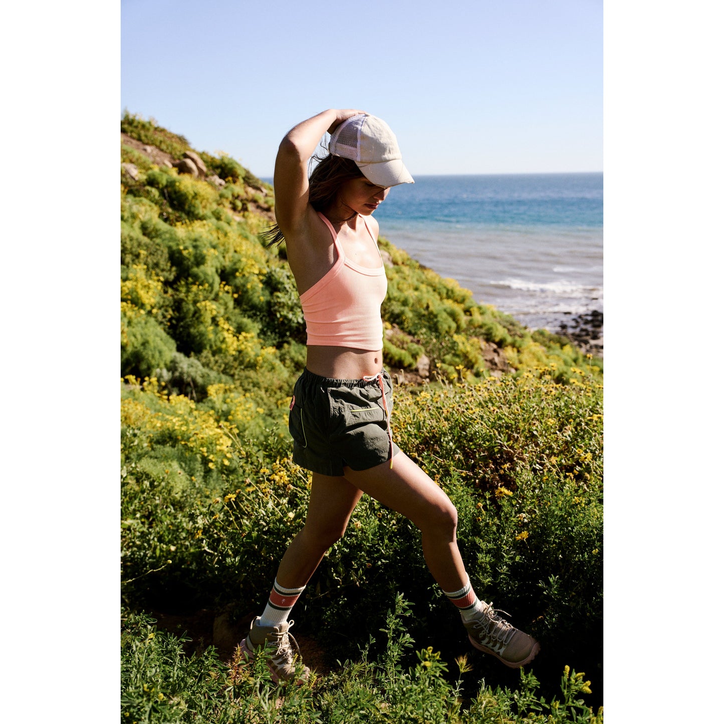 A woman in sports attire hiking through a coastal area wearing the Outskirts Skort in Olive Combo from Free People Movement, with lush greenery and the ocean in the background.