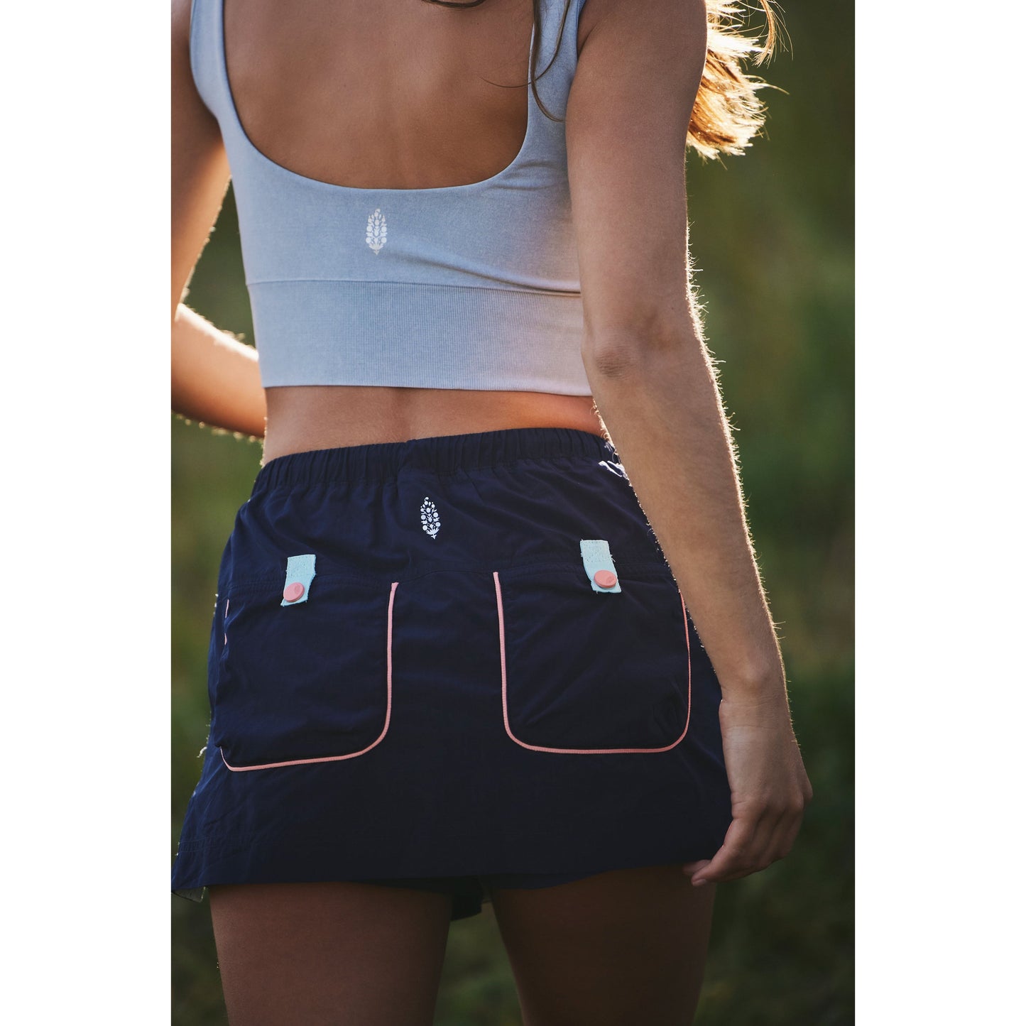 Close-up of a woman wearing a grey crop top and navy sports shorts with pink trim, standing outdoors with sunlight highlighting her Free People Movement Outskirts Skort in Midnight Navy Combo.