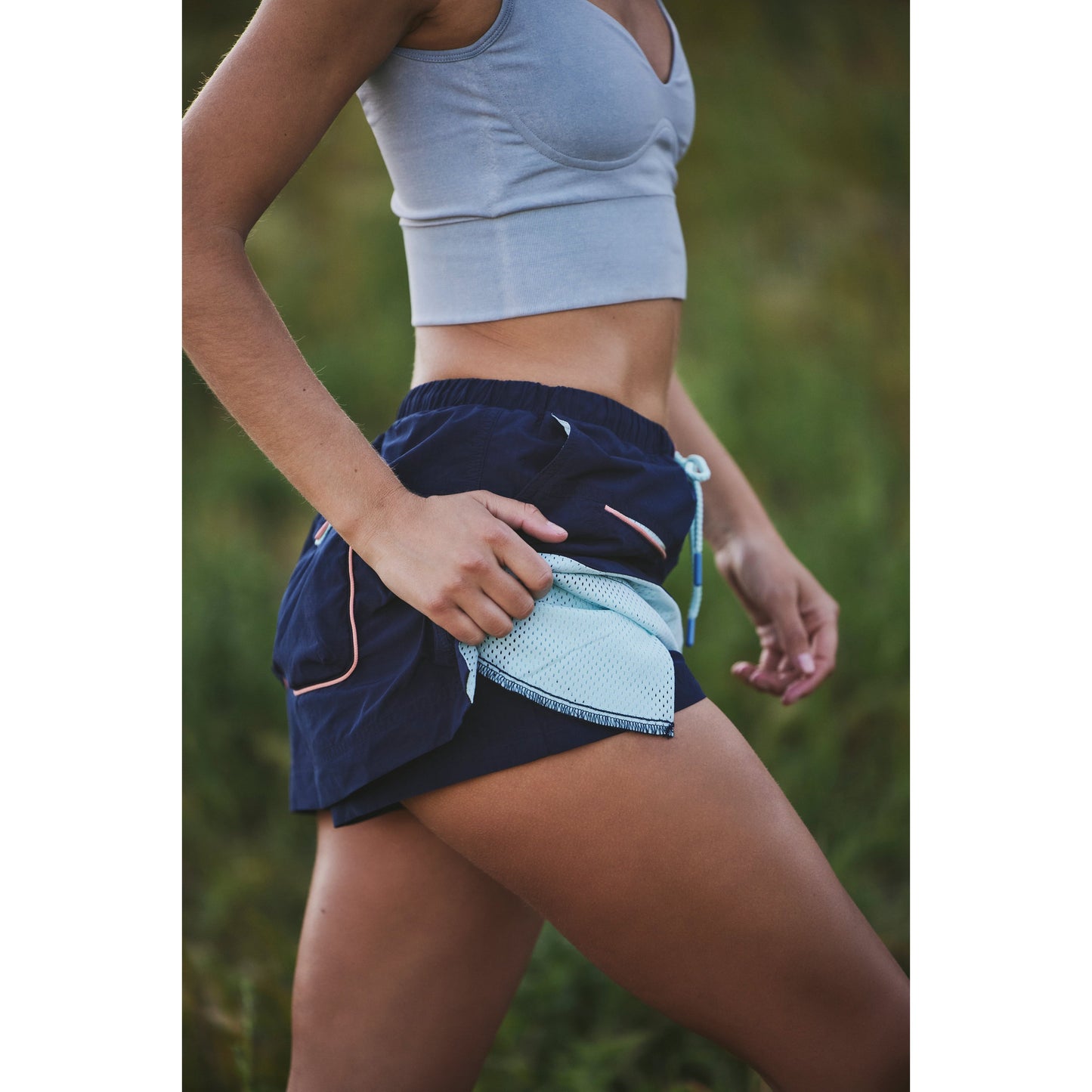 A woman runner in a gray sports bra and navy shorts putting a smartphone into a thigh pocket against a grassy background wearing the Outskirts Skort from Free People Movement.