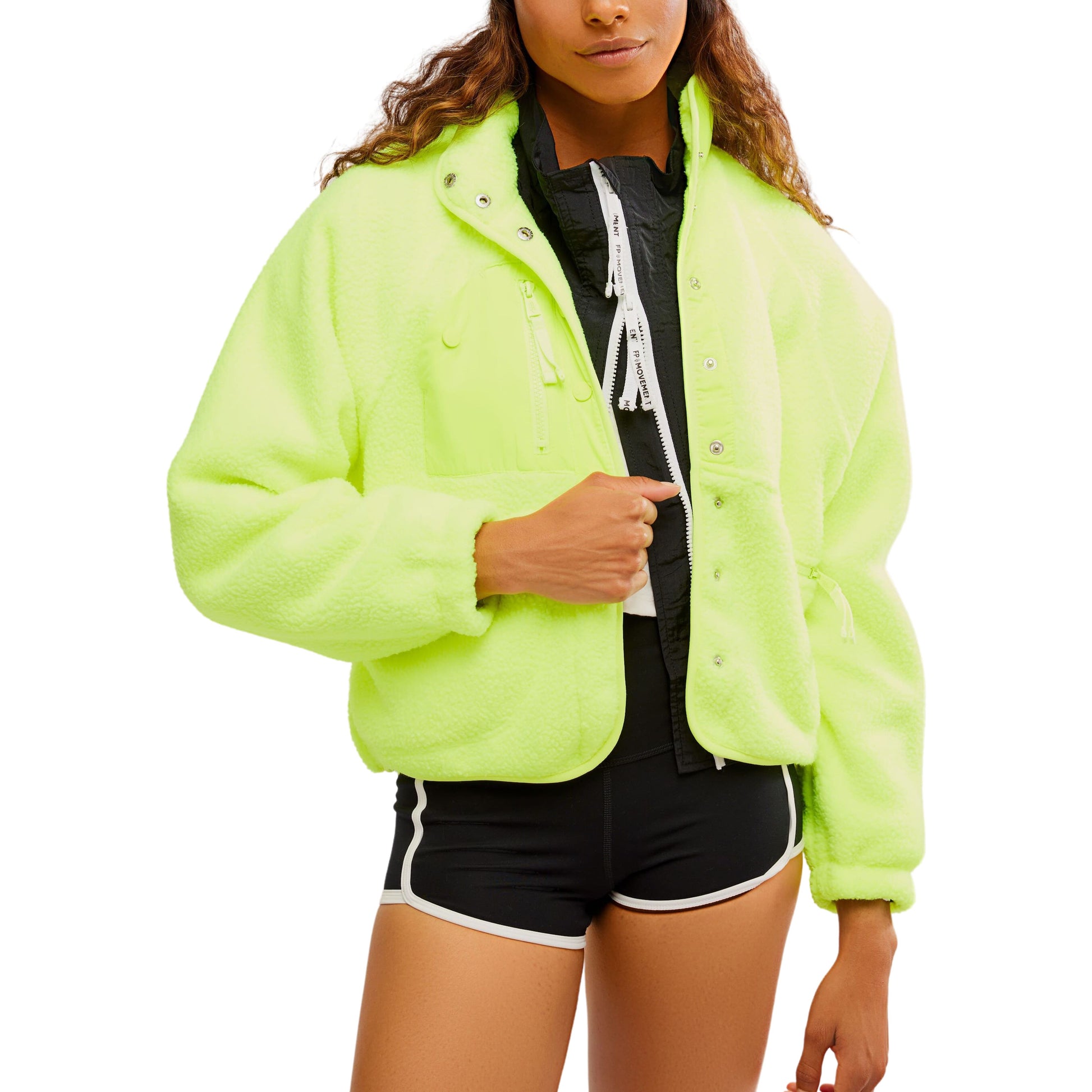 Woman in a Free People Movement Hit The Slopes Jacket in Highlighter with zippered pockets and black shorts, standing with one hand adjusting the jacket zipper.