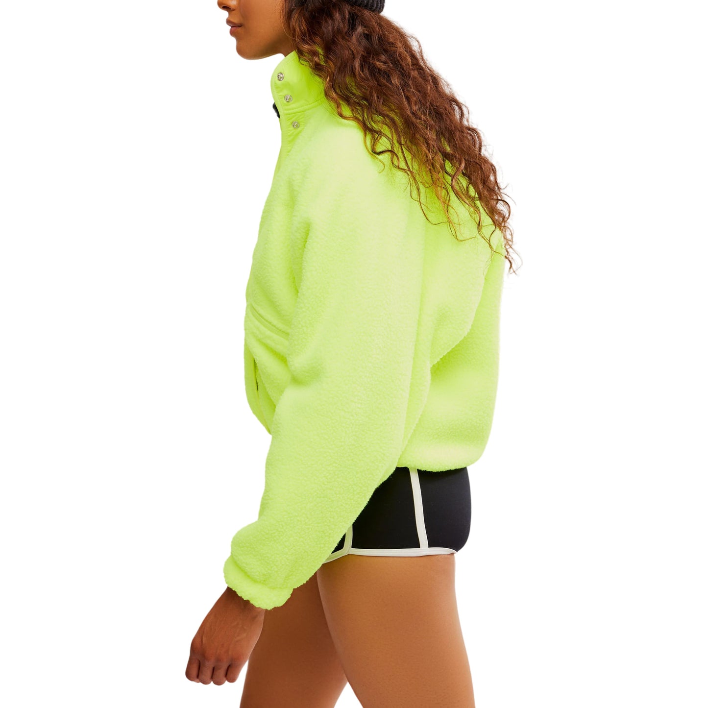 Side profile of a woman wearing a Free People Movement Hit The Slopes Jacket in Highlighter green with zippered pockets and black shorts, walking against a white background.