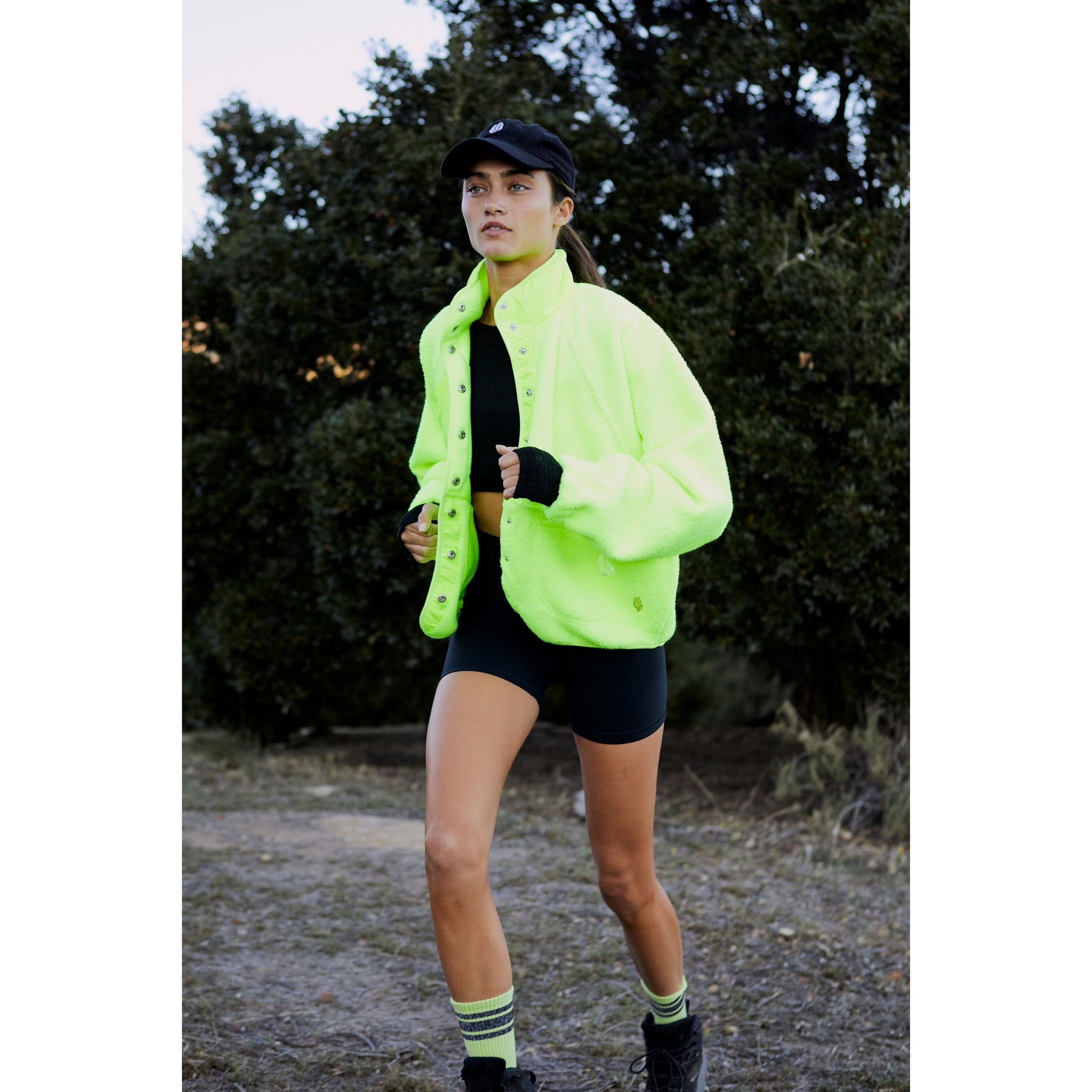 A woman in a Free People Movement Hit The Slopes Jacket in Highlighter and black shorts jogging on a trail with trees in the background.