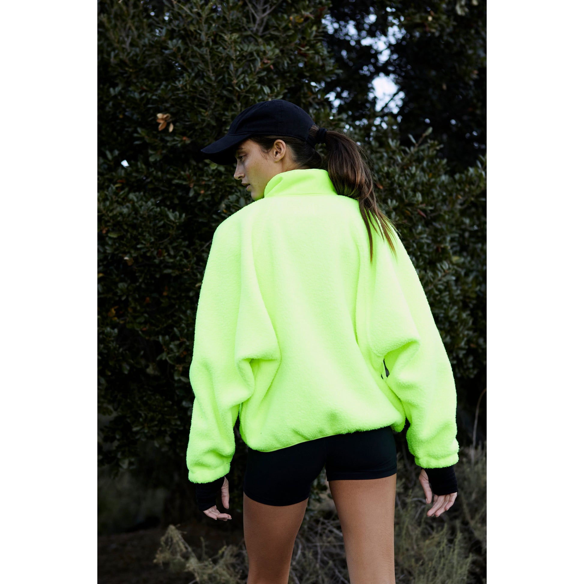 Woman in Free People Movement's Hit The Slopes Jacket in Highlighter green with zippered pockets and black shorts standing outdoors with a black cap, facing away from the camera.