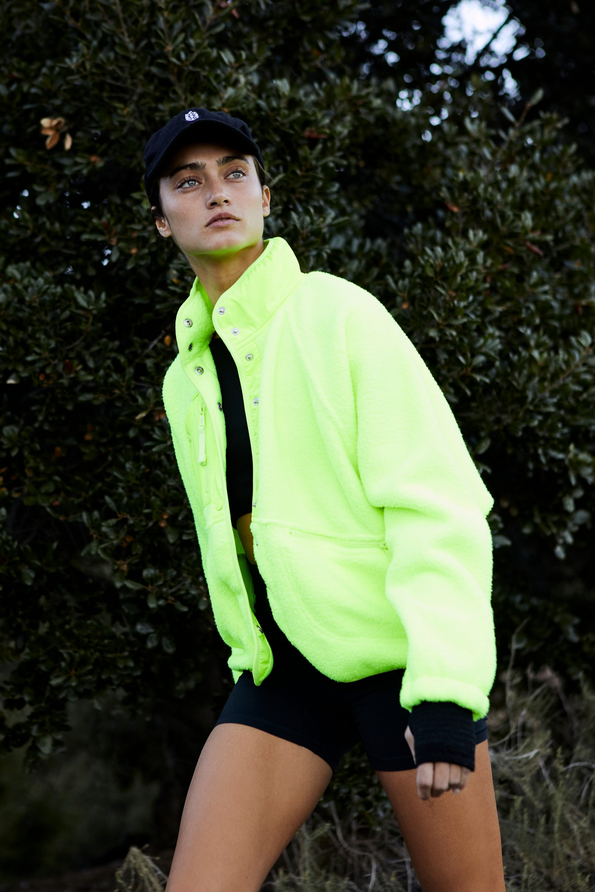 A person wearing a Free People Movement Hit The Slopes Jacket in Highlighter neon green with zippered pockets and a black cap stands outdoors with a thoughtful expression, looking away from the camera.