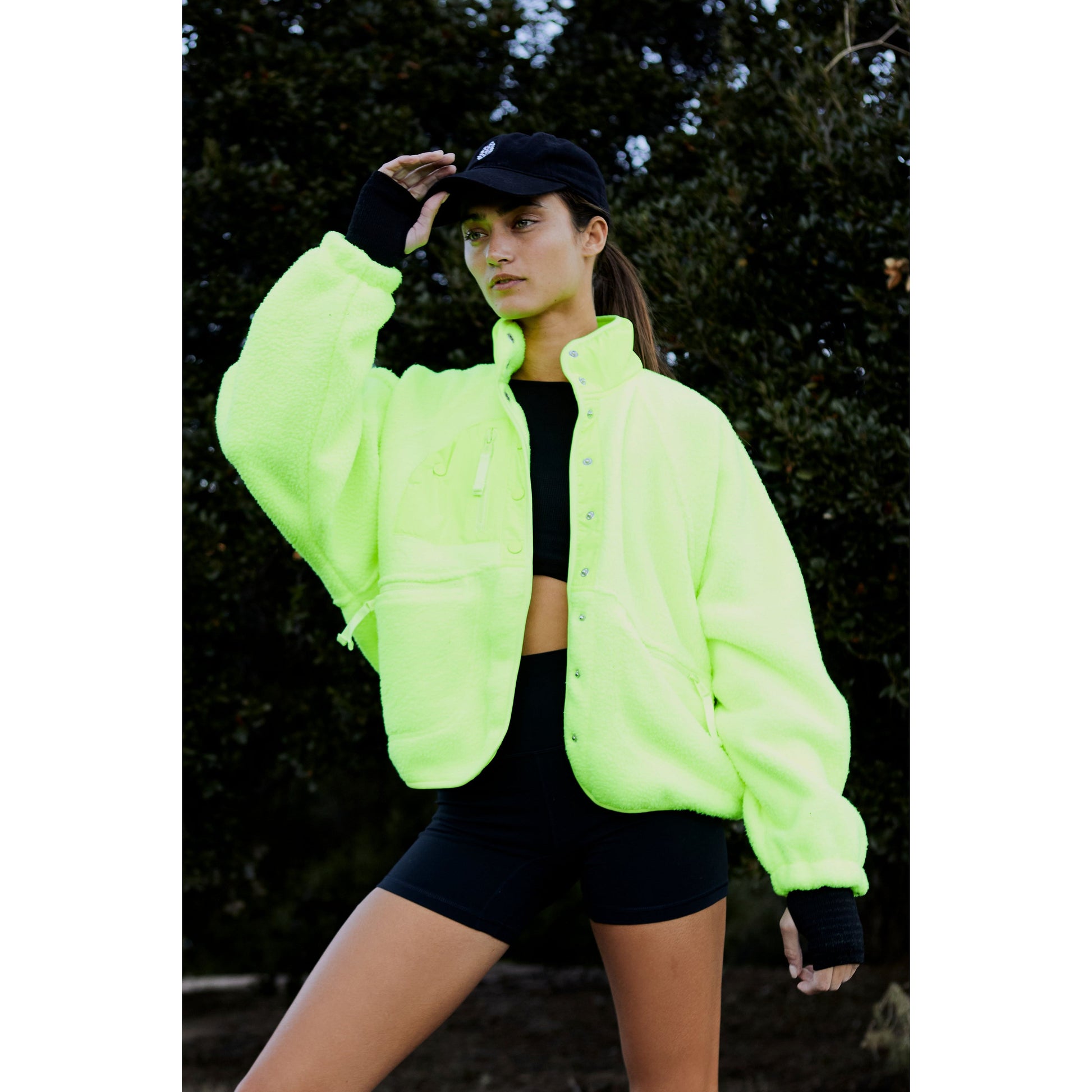 A woman in a Free People Movement Hit The Slopes Jacket in Highlighter and black cap stands outdoors, shading her eyes with one hand, with trees in the background.