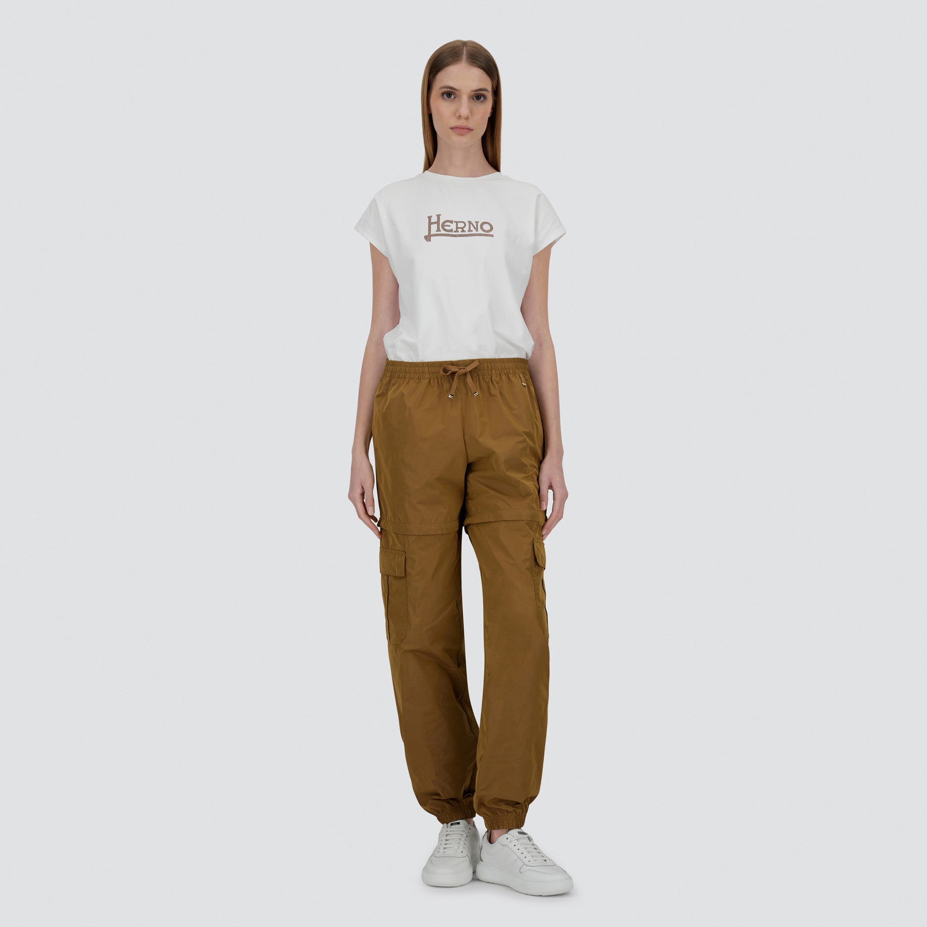 A woman faces forward against a plain light background, wearing a white Herno t-shirt, brown Herno Taffeta Cargo Zip pants, and white sneakers.