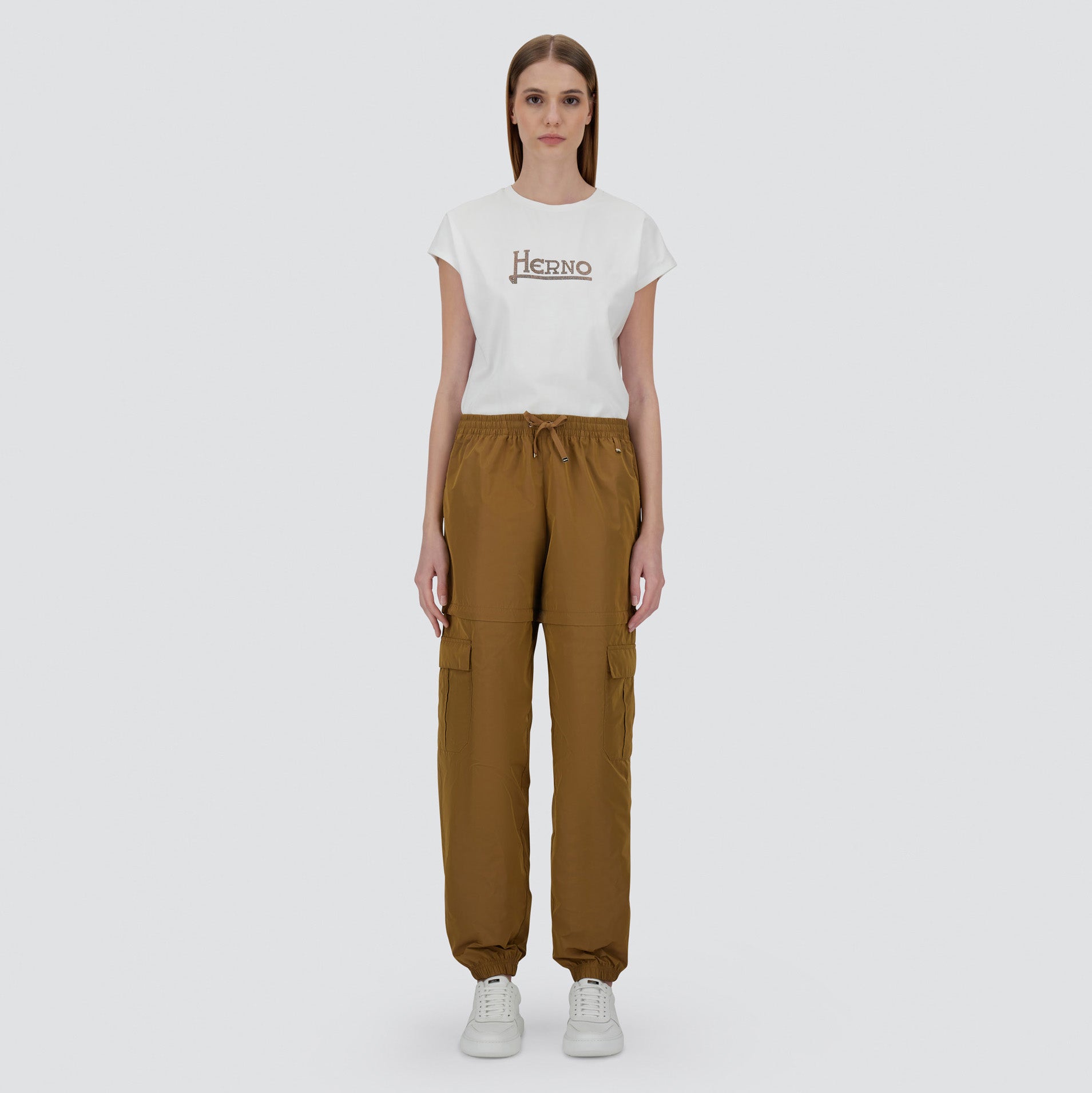 A woman faces forward, wearing a white Herno t-shirt, Taffeta Cargo Zip brown pants by Herno, and white sneakers against a plain light background.