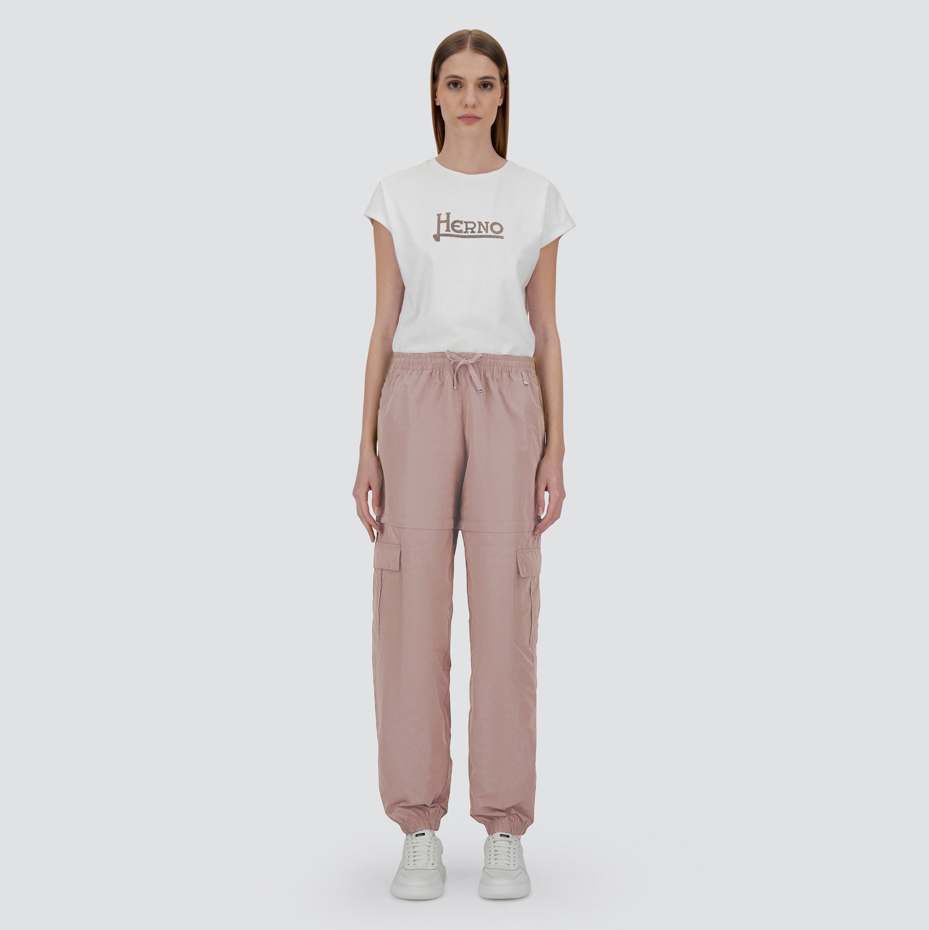 A woman faces forward in a white Herno t-shirt, light pink Herno Taffeta Cargo Zip pants, and white sneakers against a plain light backdrop.