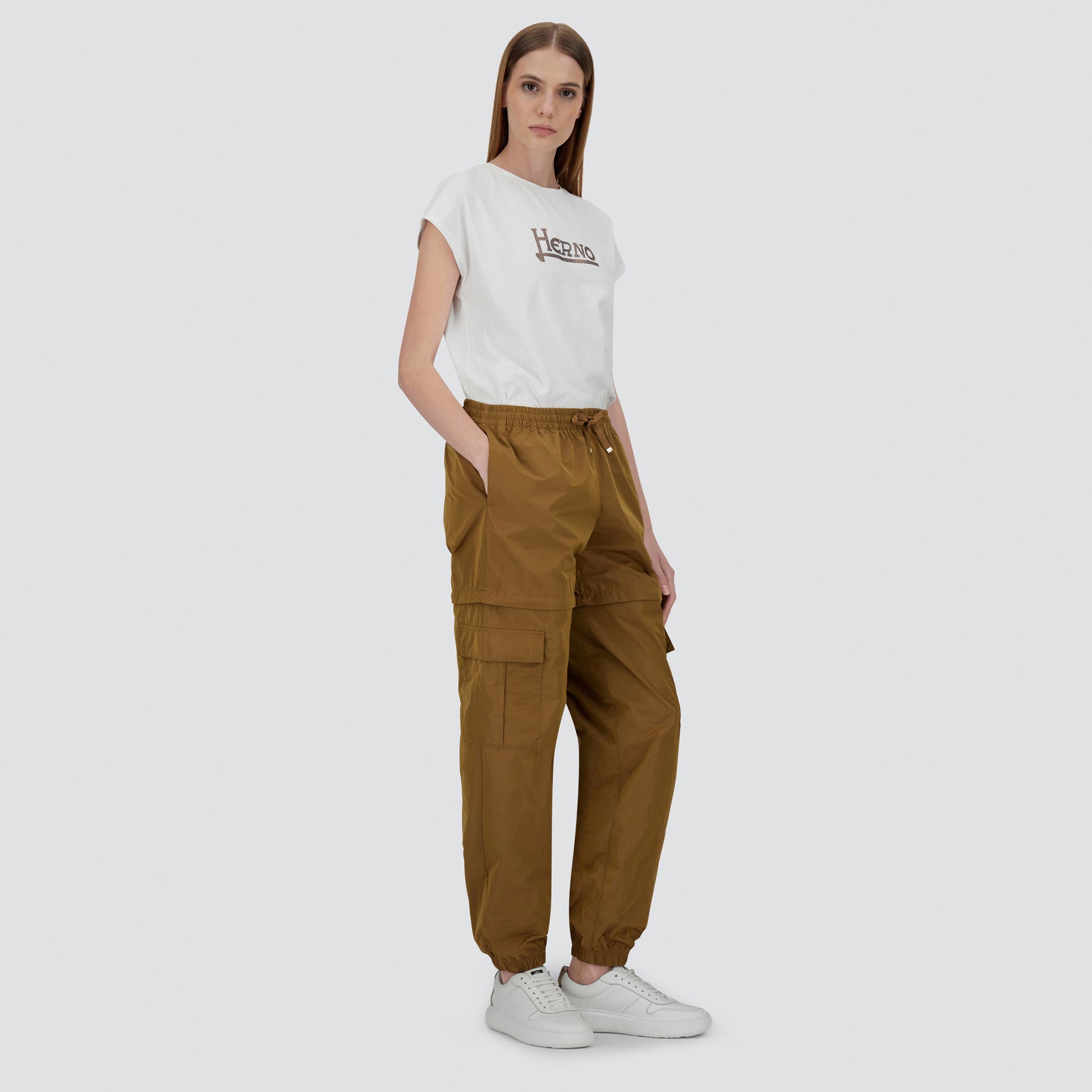 A woman poses in Herno’s Taffeta Cargo Zip brown pants with pockets, a white T-shirt with a black logo, and white sneakers against a plain background.