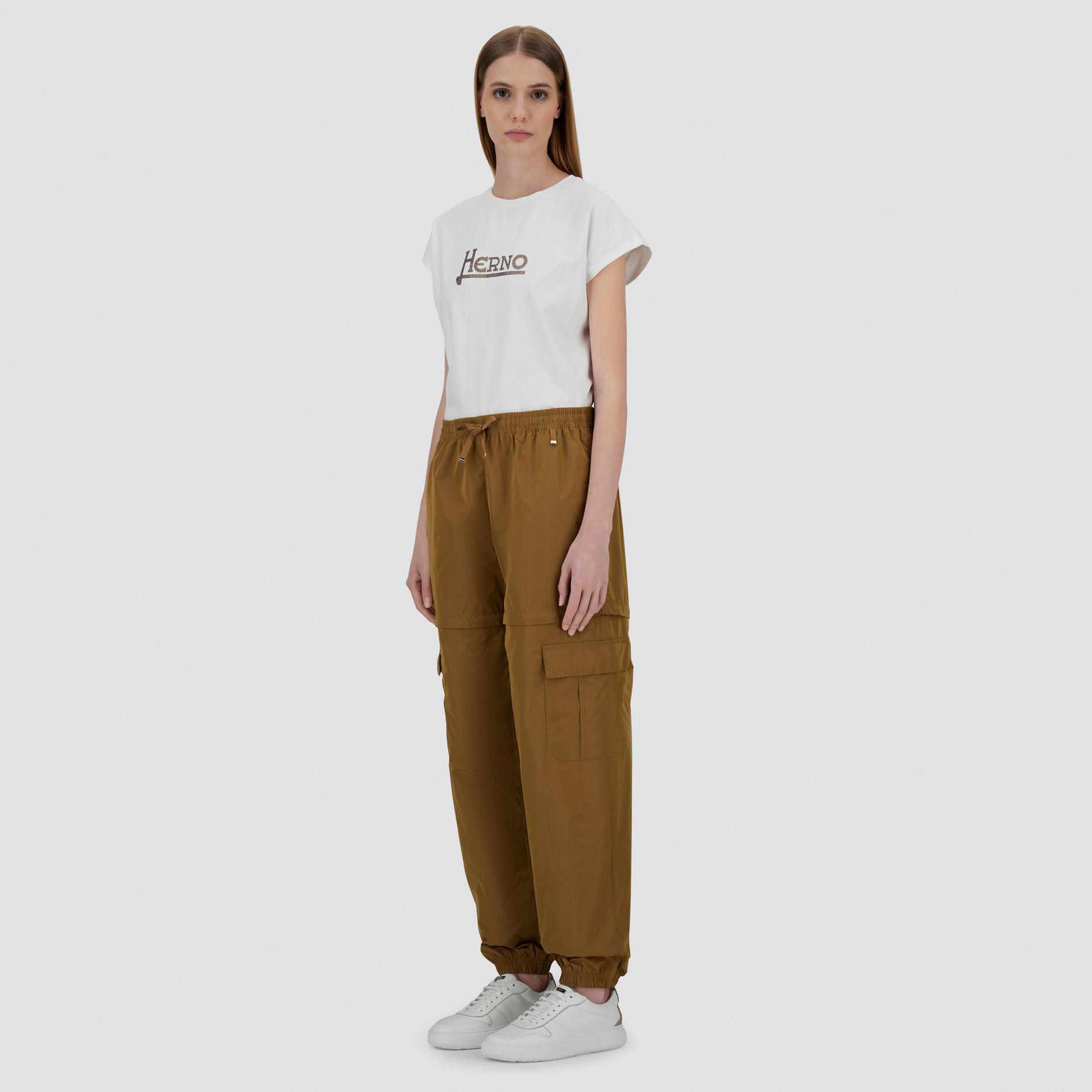 A woman faces forward against a plain light background, wearing a white Herno t-shirt, Taffeta Cargo Zip brown pants by Herno, and white sneakers.