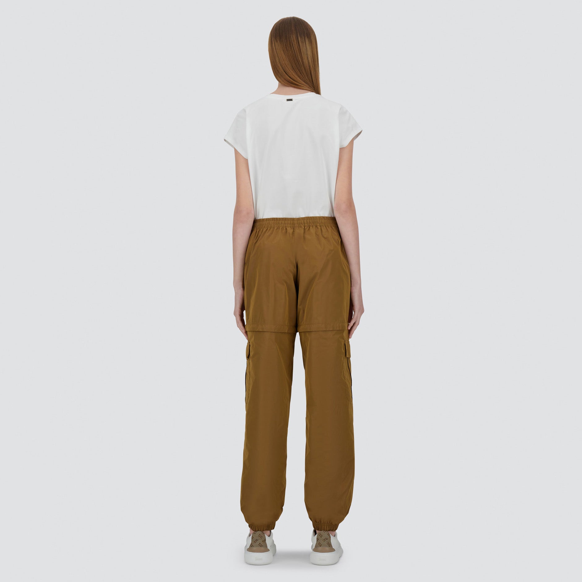 A person stands facing away, wearing Herno's Taffeta Cargo Zip brown cargo pants, a white short-sleeve shirt, and beige sneakers against a plain light background.
