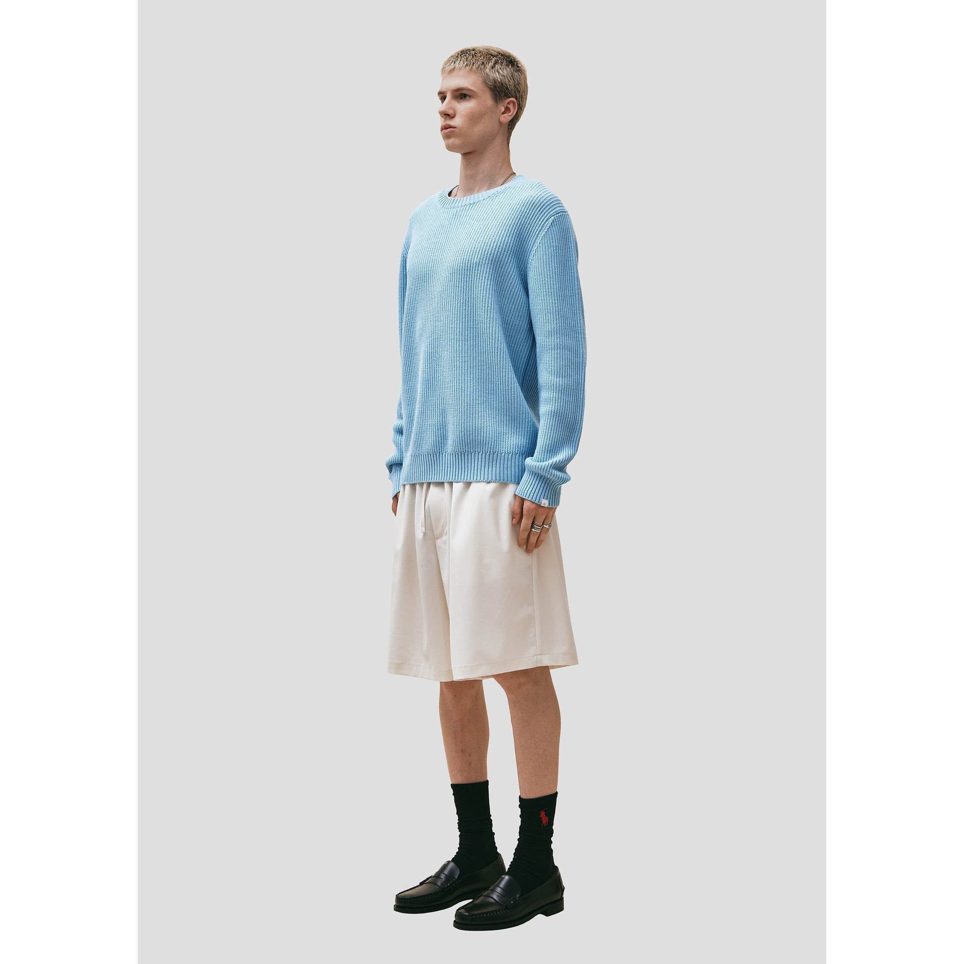 A young man standing in a studio wearing a Wool Cotton Crewneck Sweater from Seven Gauge, beige knee-length shorts, black socks, and black shoes.
