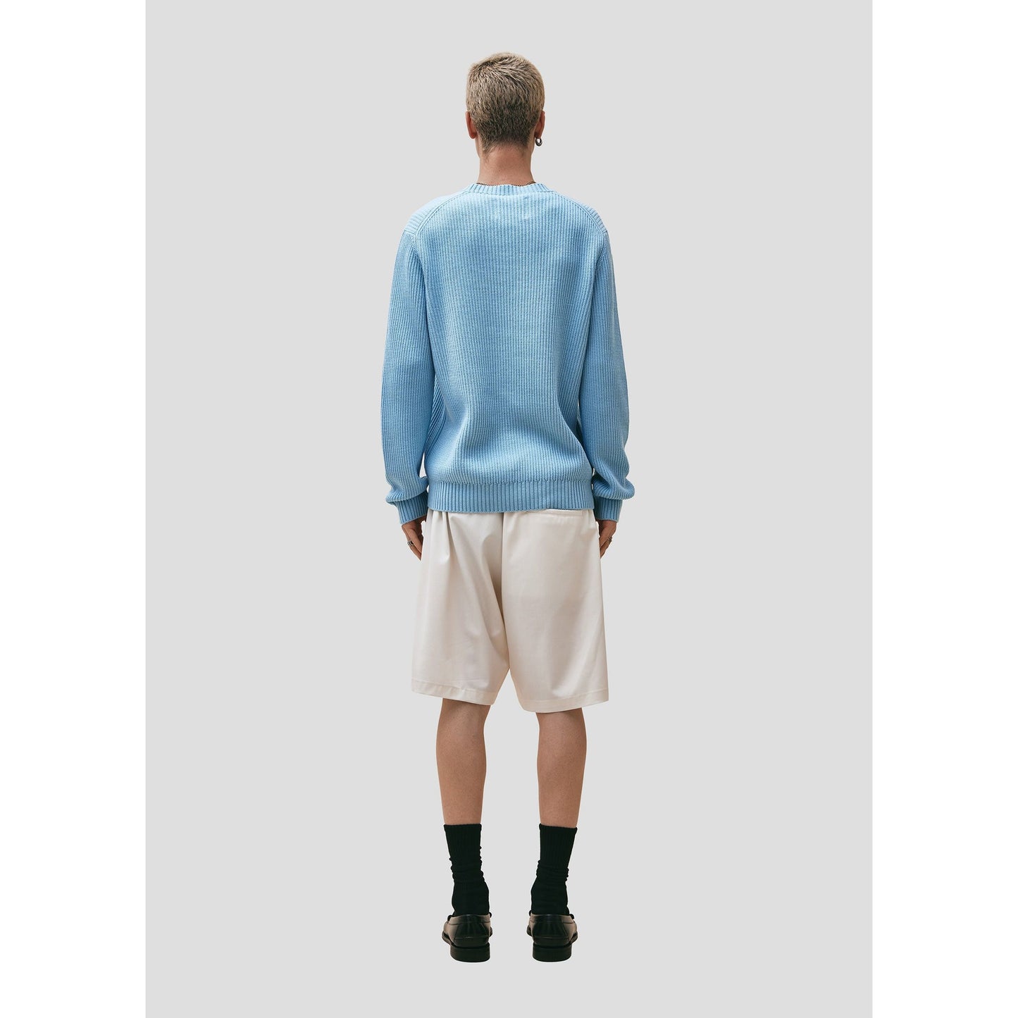 Man seen from behind wearing a Seven Gauge Wool Cotton Crewneck Sweater in Azzurro, white shorts, black socks, and brown shoes, standing against a gray background.