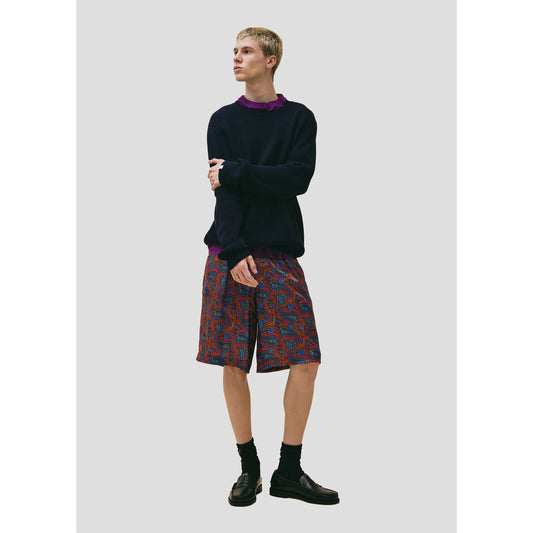 A young man stands against a light gray background wearing a dark navy Wool Cotton Crewneck Sweater from Seven Gauge, colorful patterned shorts, and black boots. He looks to the side with a neutral expression.
