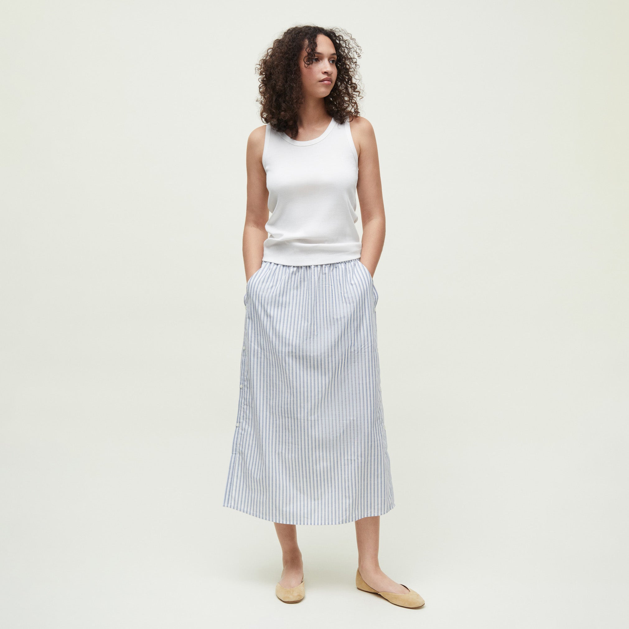 A woman faces forward against a plain background, wearing the Aiayu Rib Tank in white, paired with a light blue and white striped midi skirt and beige flats, her hands tucked into her skirt pockets.
