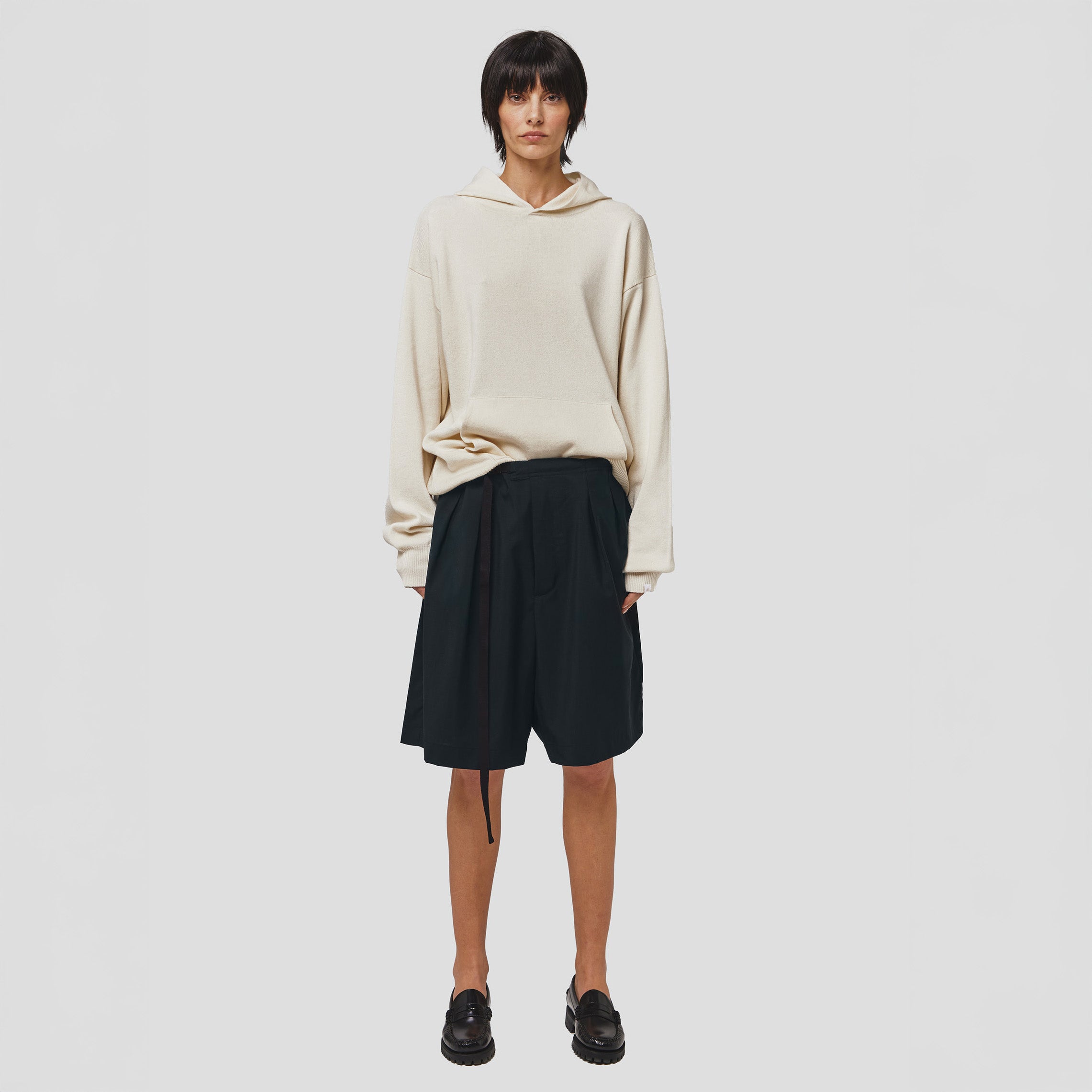A person stands against a plain white background, wearing a cream hoodie, Seven Gauge Tropical Wool Basket Shorts with an adjustable waist strap, and black loafers.
