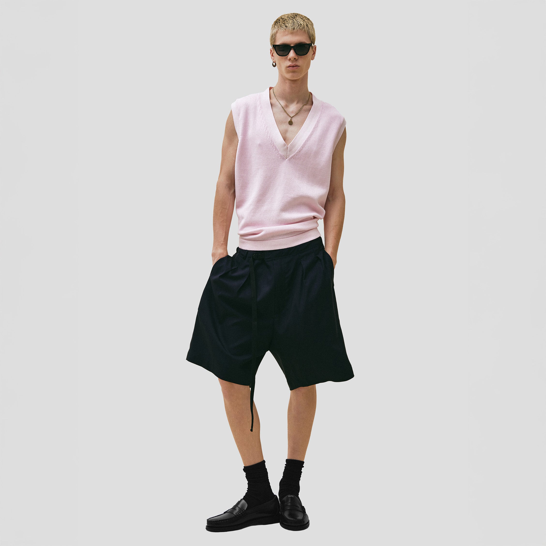 Wearing a pink sleeveless V-neck top, sunglasses, and black shoes, a person stands against a plain background dressed in Seven Gauge's Tropical Wool Basket Short with an adjustable waist strap.
