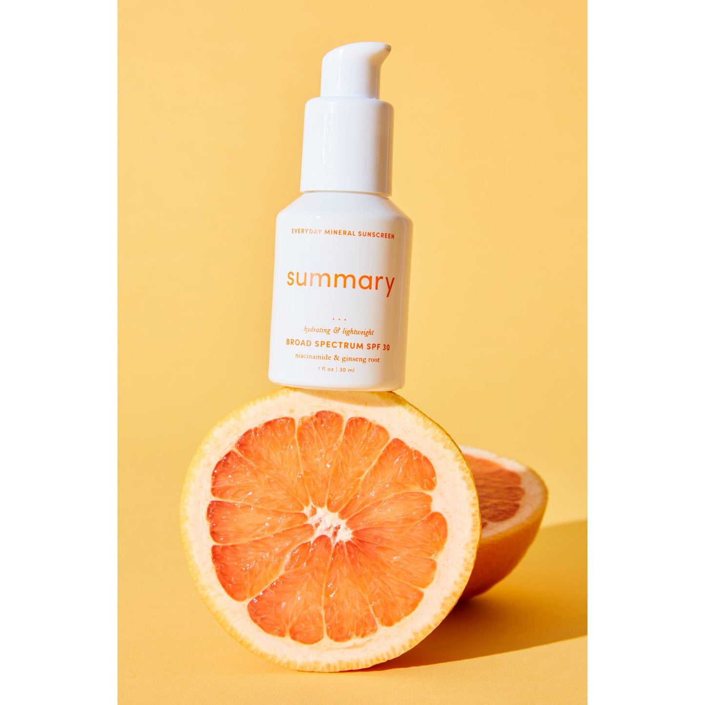 A bottle of "Free People Movement" brand Summary SPF 30 broad spectrum mineral-based sunscreen placed behind a sliced grapefruit, set against a yellow background.