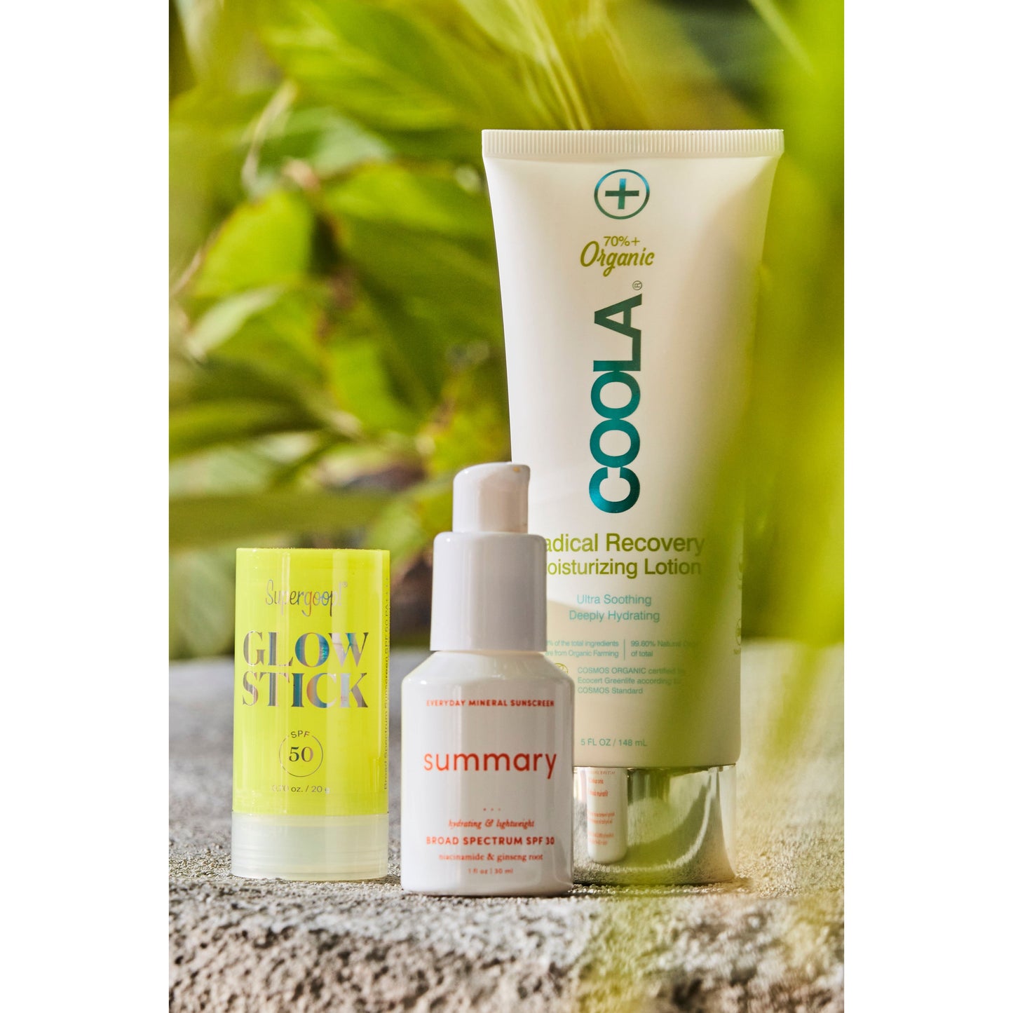 Three skincare products by Coola and Supergoop, including a mineral-based sunscreen, displayed on a natural stone surface, surrounded by lush greenery.