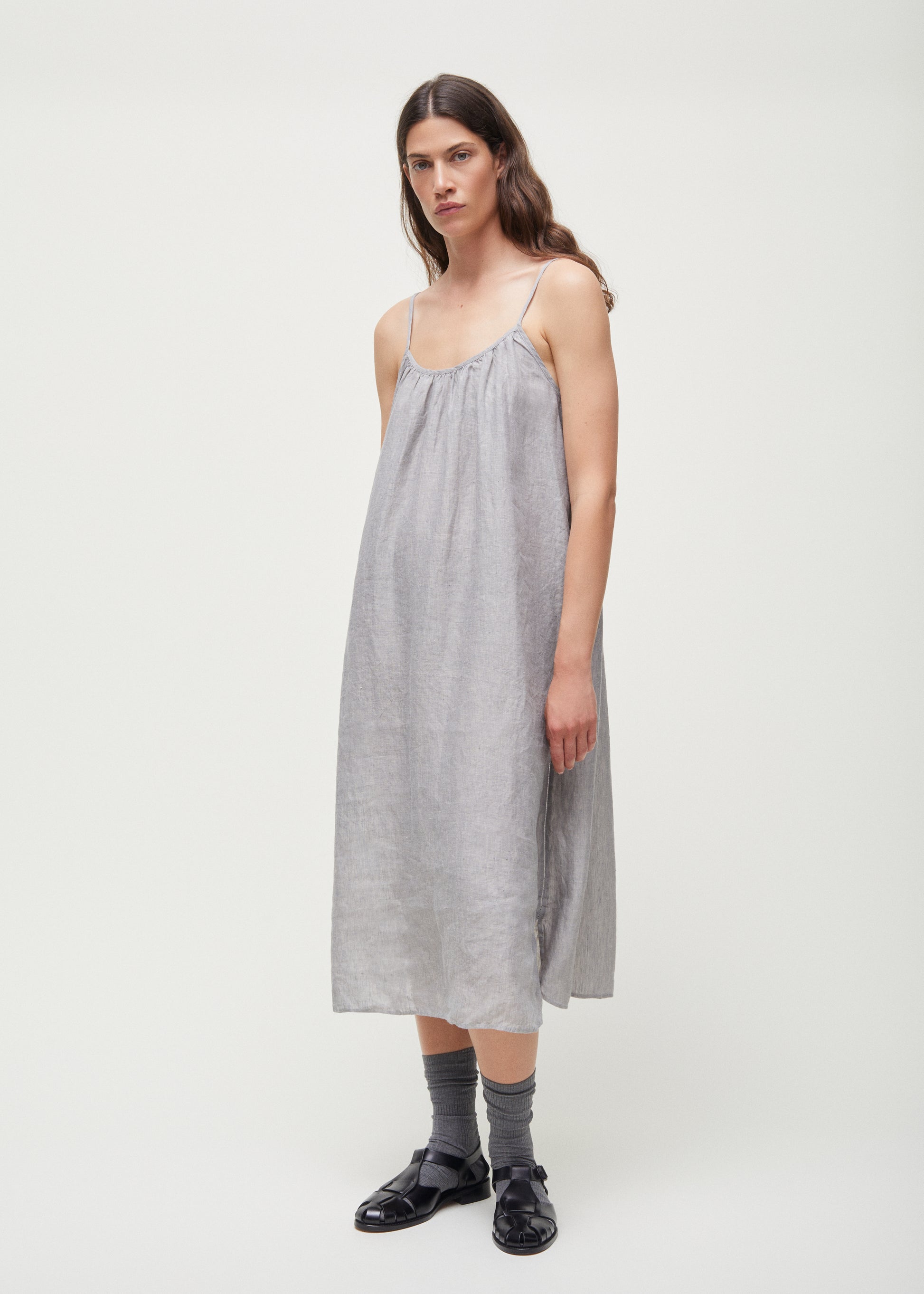 A person stands against a plain white background wearing the Aiayu Strap Dress Linen, featuring loose-fitting European linen with thin straps, complemented by gray socks and black sandals.